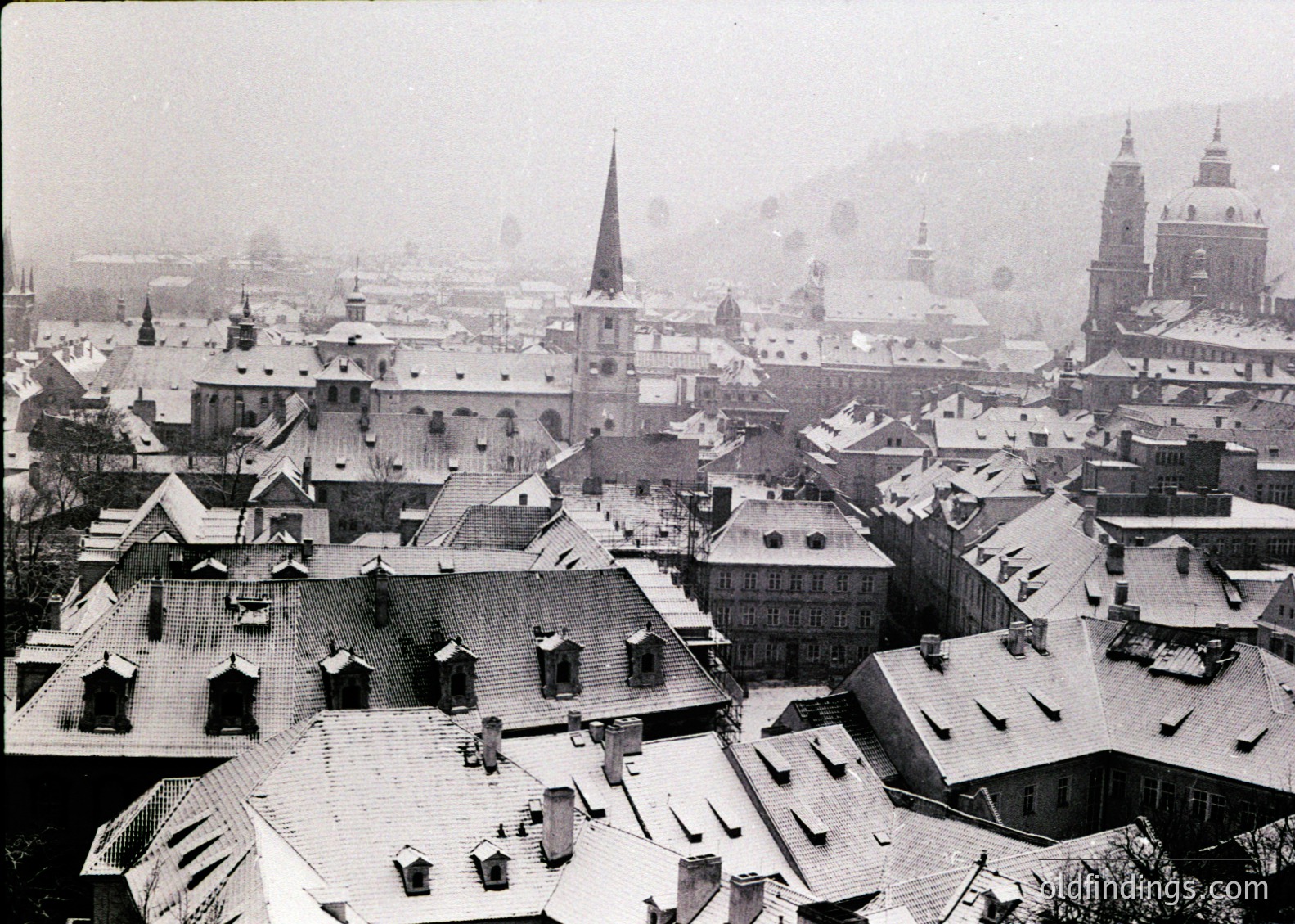 Aerial view of a historic European cityscape blanketed in snow, showcasing dense, low-rise architecture with steep slate roofs and chimneys. Prominent church spires and domed towers dominate the skyline, indicating a central European setting. Likely early-to-mid 20th century based on architectural style and monochrome tone.