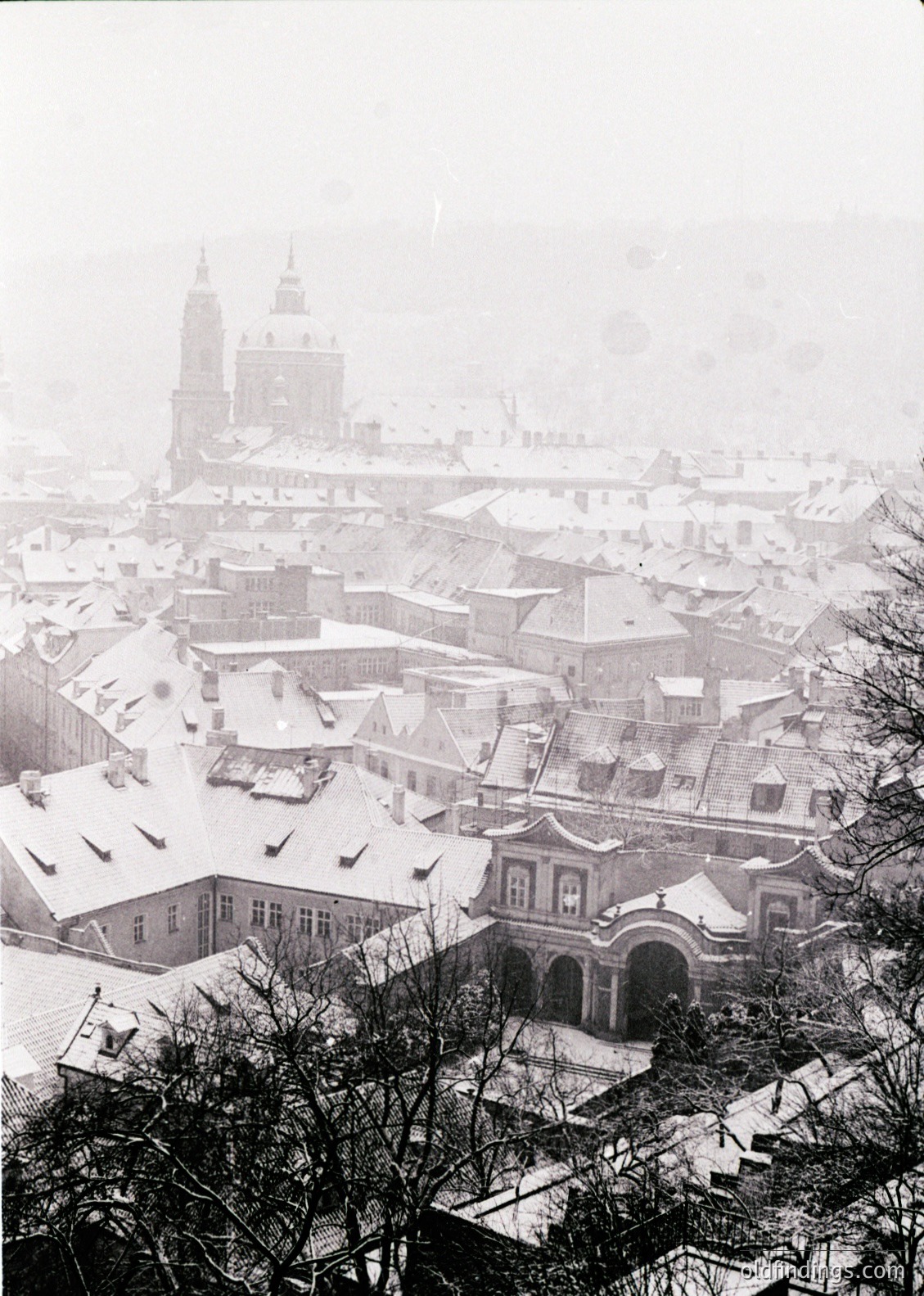 Snow-covered rooftops and historic architecture in a European cityscape, likely . Prominent Baroque-style buildings and a cathedral with twin towers dominate the skyline. Fog obscures distant details, emphasizing winter atmosphere. *(199/200 chars)*