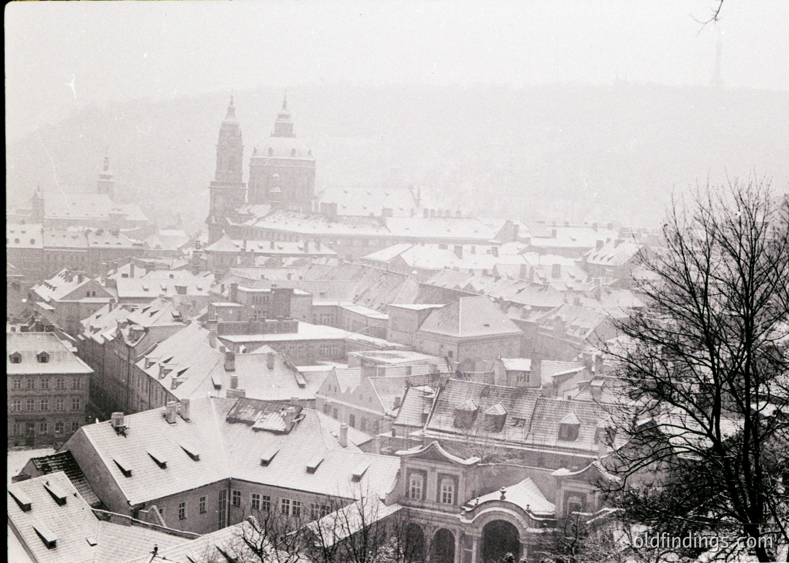 Snow-covered European cityscape with historic architecture, likely early-to-mid 20th century. Prominent domed cathedral and clustered rooftops under misty skies. Architectural style suggests Eastern European influence.