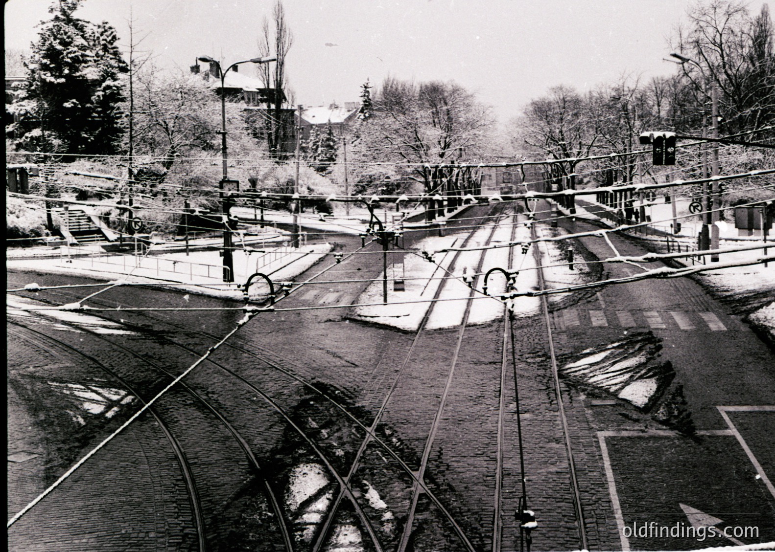 A vintage aerial view of a tram network intersecting with a canal in an urban setting. Tram tracks converge at a central junction, with overhead wires and poles. Snow covers ground and rooftops, indicating winter. Leafless trees and sparse pedestrians suggest early-to-mid 20th century. Likely European city with industrial-era infrastructure.