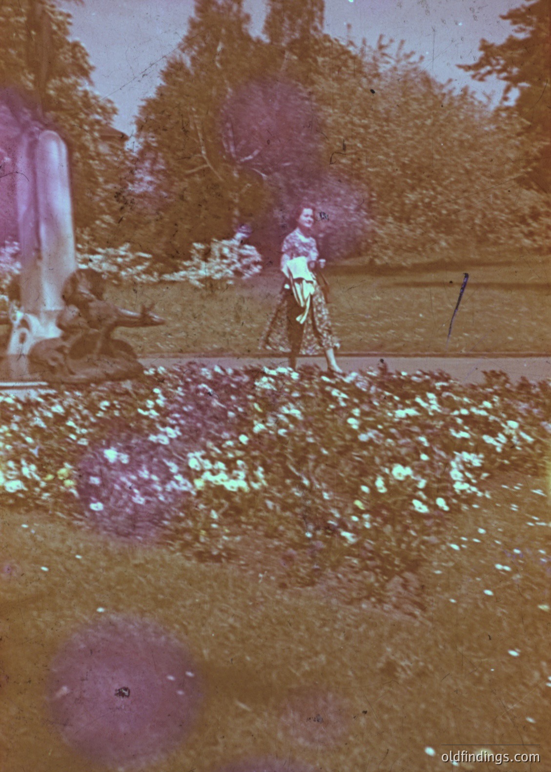 Vintage sepia-toned photograph of a lone figure in a wide-brimmed hat and patterned dress, walking on a dirt path surrounded by dense foliage. The scene suggests a rural or park setting, likely late 19th to early 20th century. The aged paper and fading tones highlight historical photography techniques.