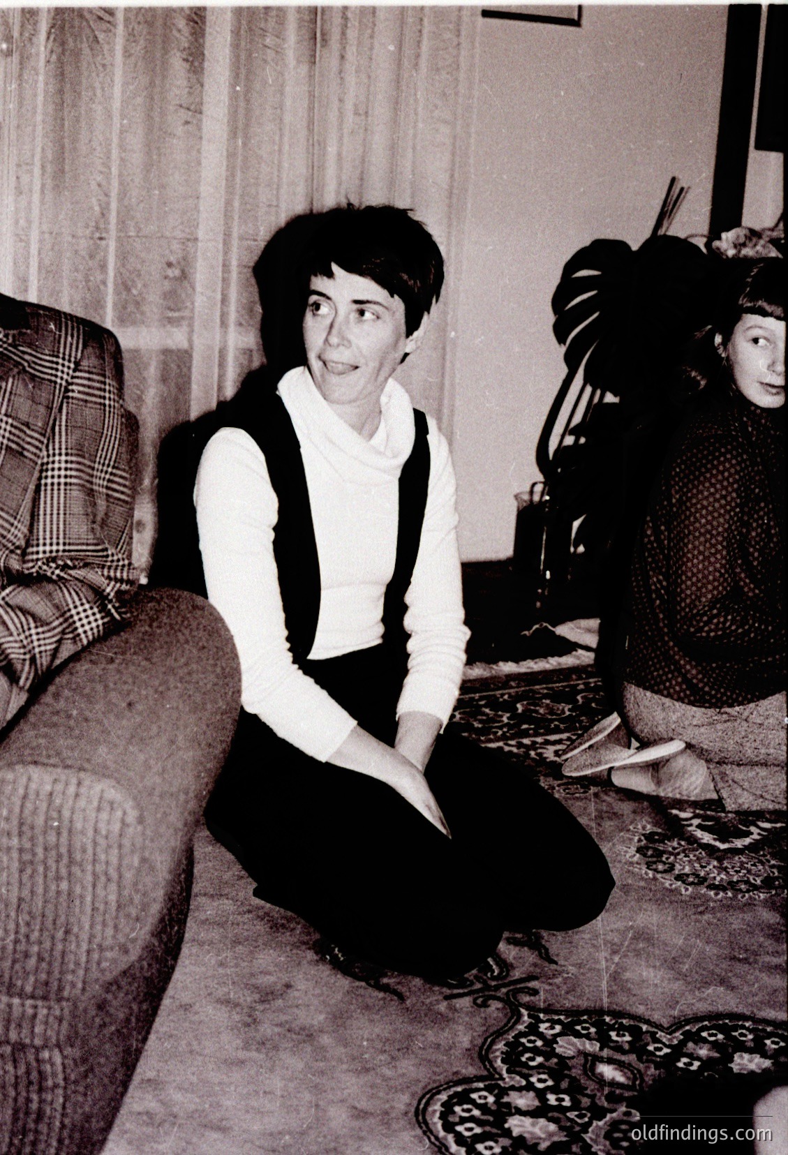 Vintage black-and-white interior shot of a woman in a cropped sweater vest and high-waisted trousers, seated on a patterned rug beside a plaid-curtained window. Partial view of a child in a knitted sweater and hat in foreground. Mid-century domestic setting, likely 1960s–1970s.