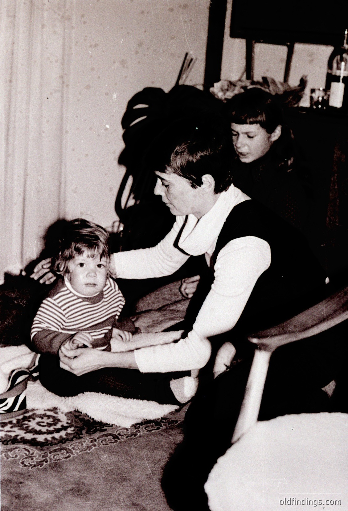 Vintage black-and-white photo of an indoor domestic scene, likely mid-20th century. A woman in a striped sweater and apron kneels beside a young child on a patterned rug, while another woman stands in the background. Wooden furniture and a floral-patterned wallpaper suggest a mid-century household setting.