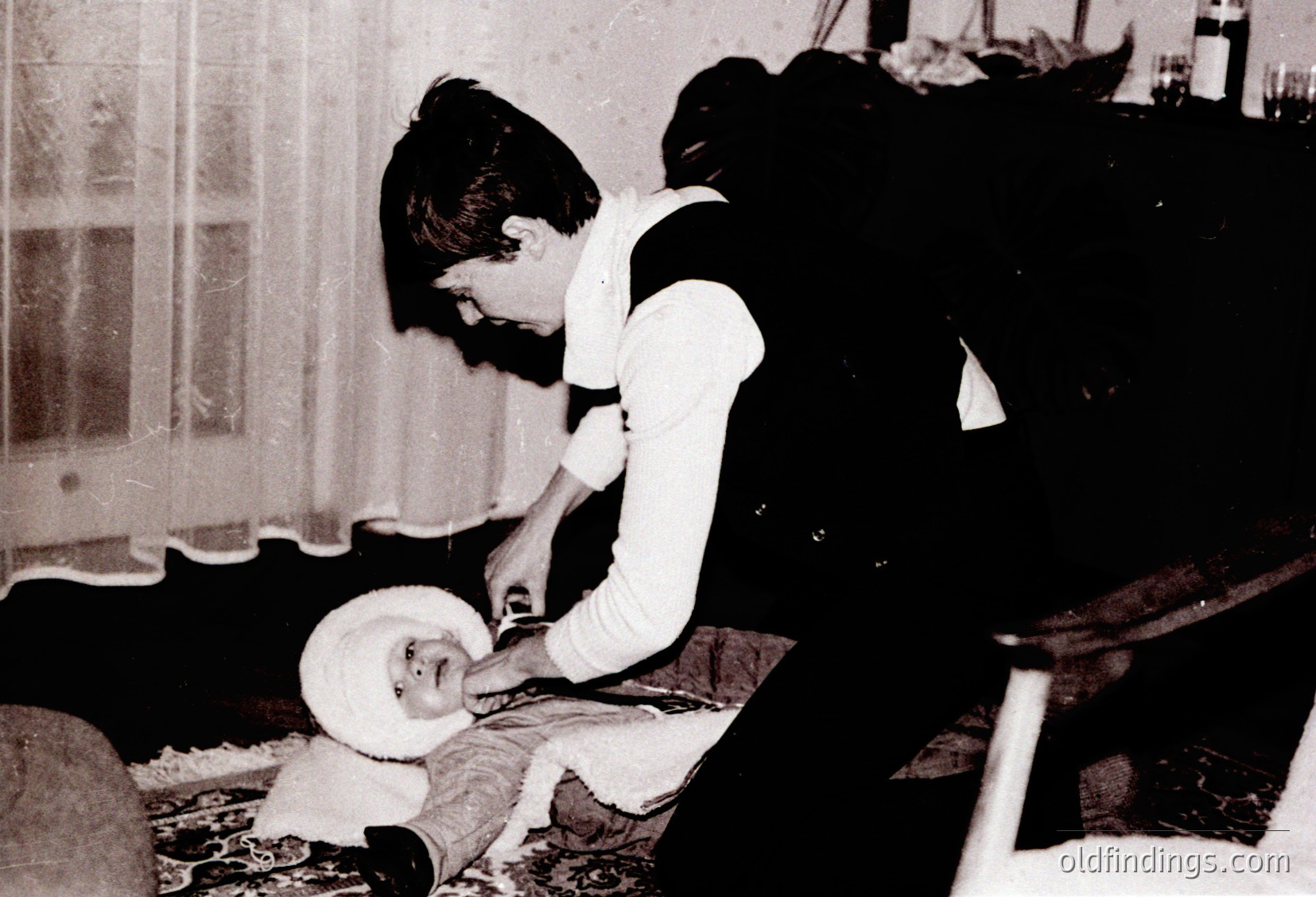 Mid-20th century indoor scene: a woman in a white nurse’s uniform adjusts a baby’s clothing on a wooden chair. Plain curtains and a simple table with crockery suggest a modest home setting.