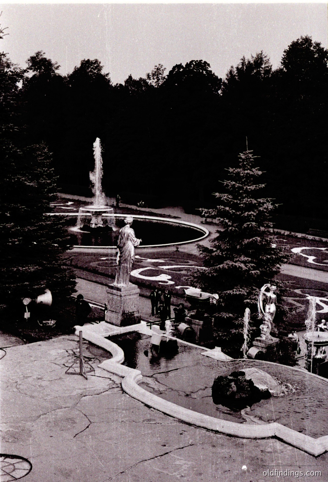 Symmetrical Baroque-style fountain with central water spout and sculpted figures in a sunken circular basin, surrounded by curved stone pathways. Lush evergreen trees frame the scene, suggesting a European garden from the 17th–18th century.
