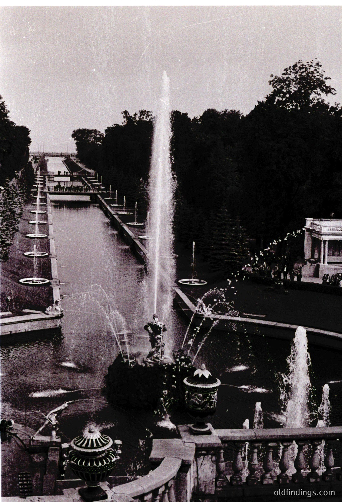 Symmetrical canal flanked by tiered fountains and ornate planters, likely part of a grand European palace or park. Mid-20th century (1950s–1960s) architectural landscaping with classical influences.