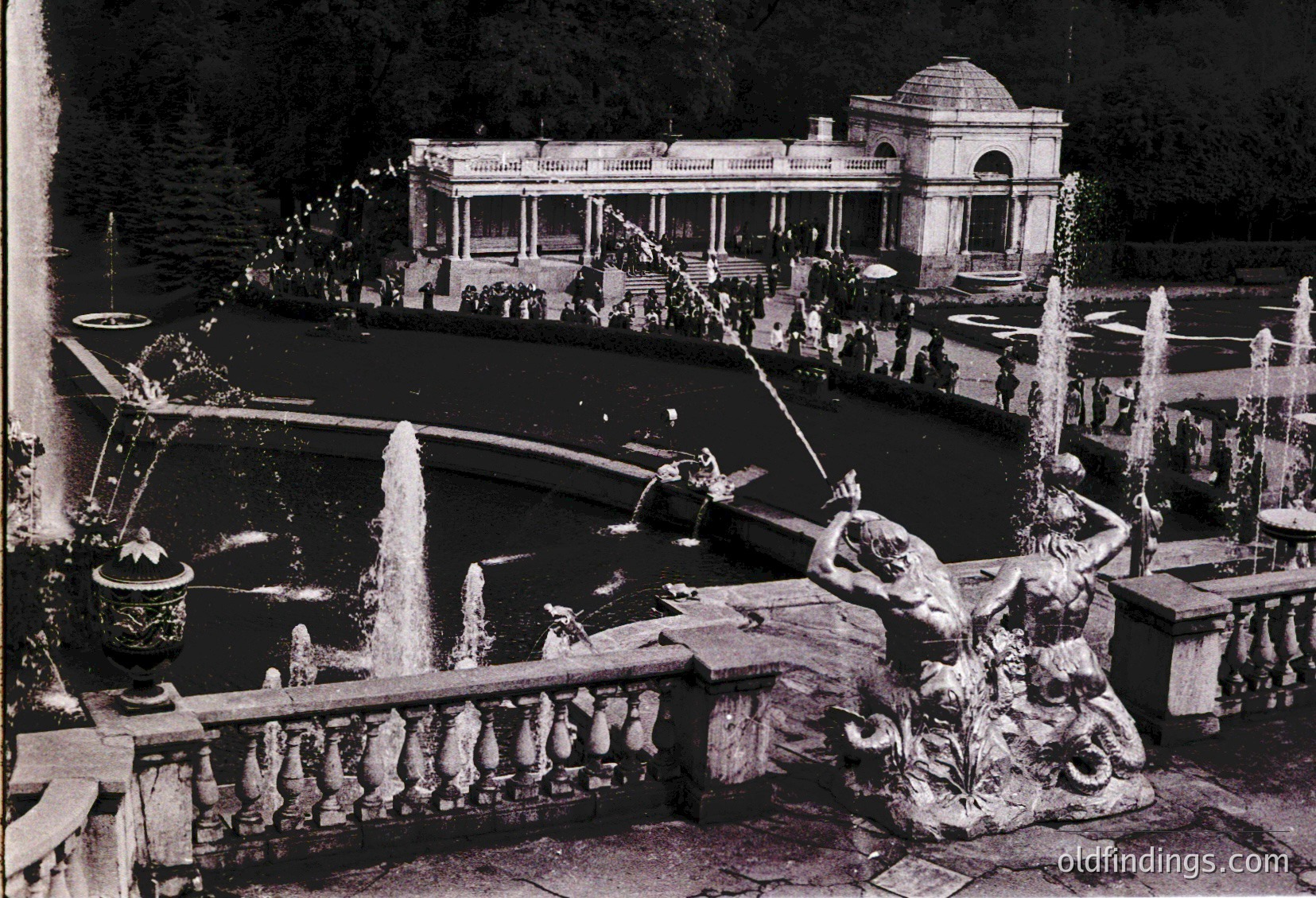 Vintage black-and-white photo of an ornate **fountain courtyard** with classical sculptures and cascading water. Central **triton statue** sprays water into a pool, flanked by decorative urns and balustrades. Background features a **pavilion-style building** with arched windows and a domed roof, surrounded by lush greenery and a crowd of people.