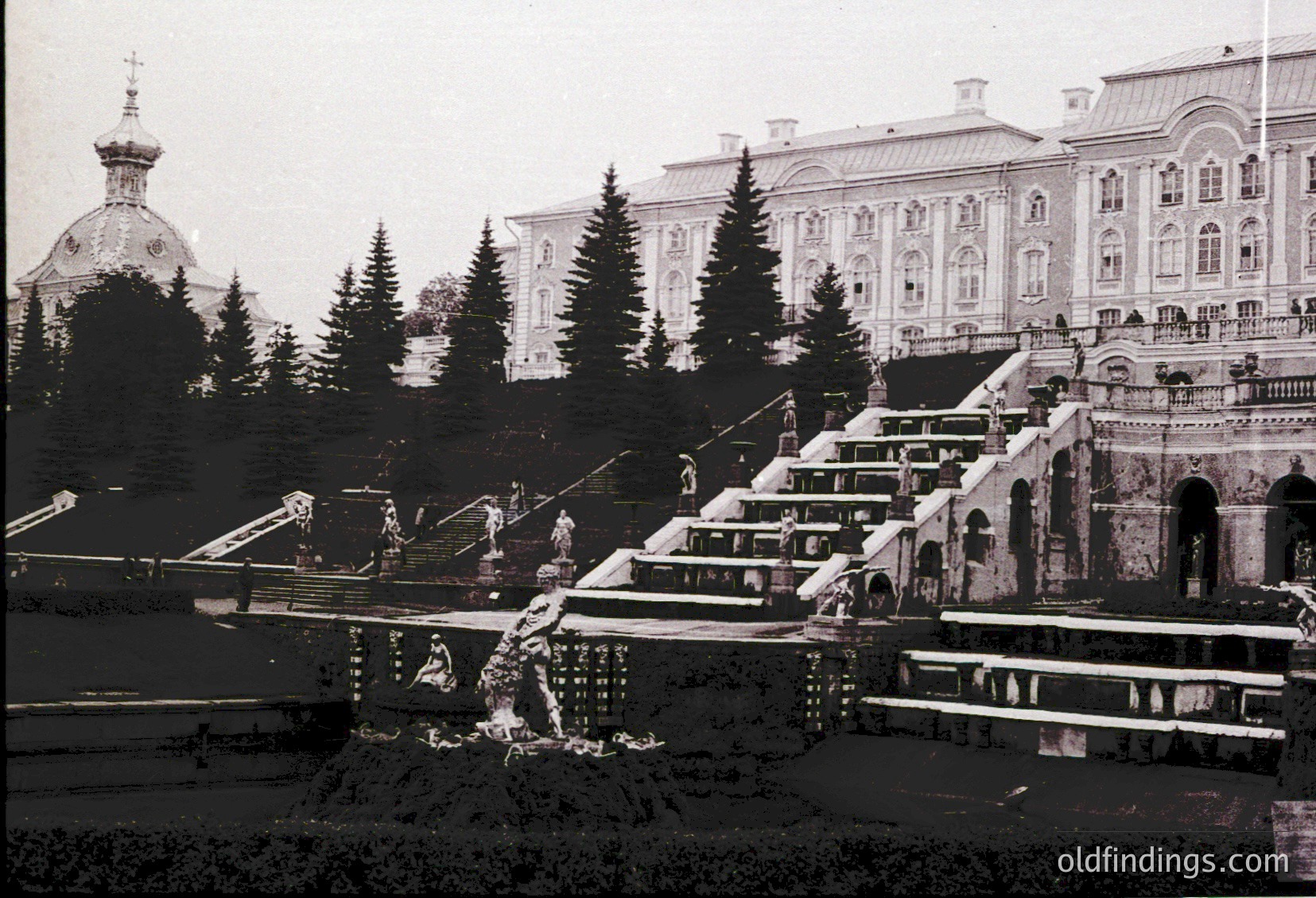 Vintage black-and-white view of grand **staircase cascading into tiered pools**, flanked by classical architecture and coniferous trees. Prominent **domed church** with ornate spire in background. Likely **early 20th-century European palace grounds**, possibly **Russia or Eastern Europe**.