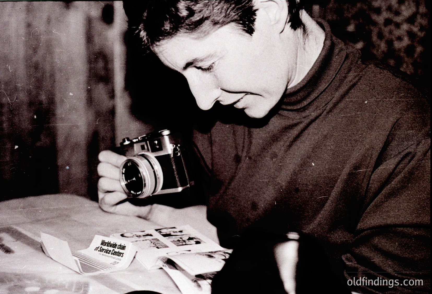Vintage black-and-white portrait of an individual examining a stack of photographs using a vintage 35mm camera. The scene suggests a focus on analog photography, likely from the mid-20th century. The visible text on a wrapper hints at a wartime or post-war context ().