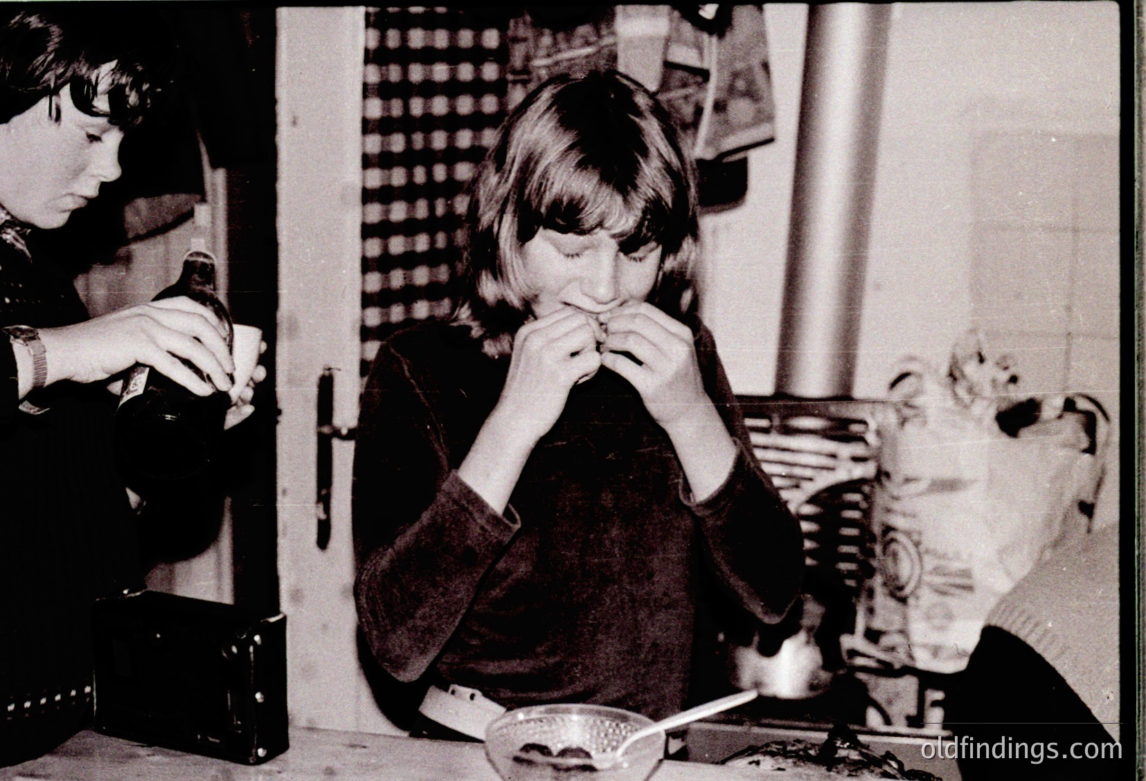 Black-and-white snapshot of two women in a domestic setting, likely 1970s–1980s. One woman sits at a table holding a spoon to her mouth, while the other stands, holding a small object. A cat rests on a chair beside a woven basket. Plain interior with a window grid pattern. Evokes vintage home life.