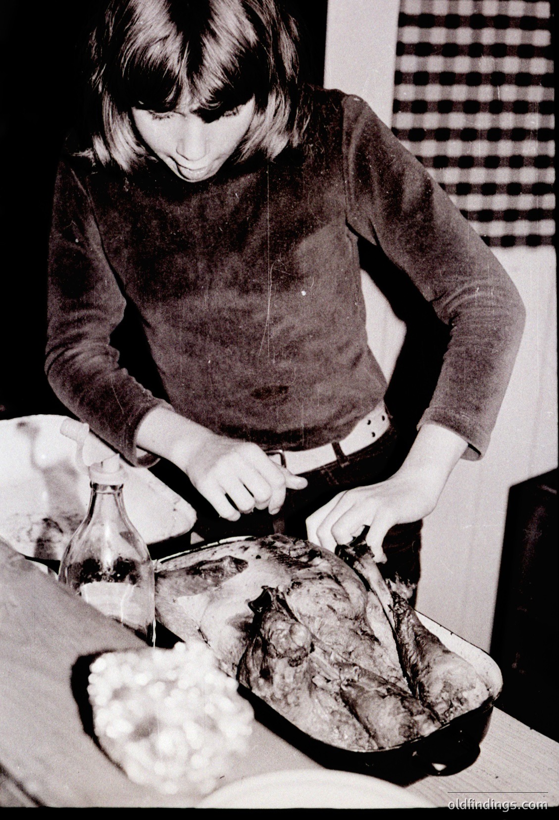 Mid-century kitchen scene featuring a woman basting a roasted turkey with a baster, surrounded by a glass bottle (likely for gravy) and a bowl of mashed potatoes. Her 1960s-style sweater and belt detail suggest domestic life in Western households. Classic holiday or family meal preparation.