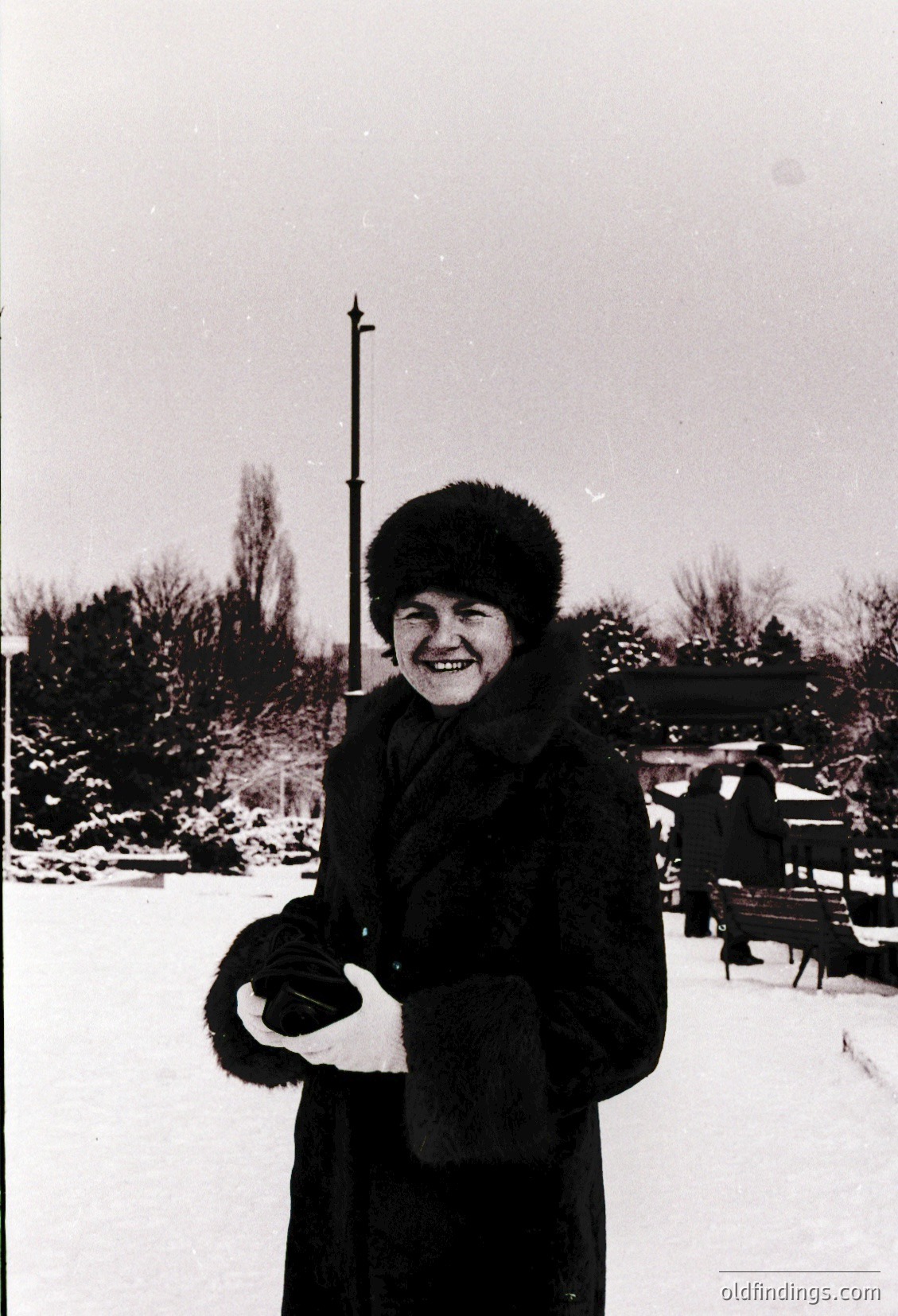 Black-and-white winter portrait of a woman in a fur-lined coat and hat, holding a fur-trimmed glove. Snow-covered park bench and leafless trees in background suggest mid-20th century urban setting. Warm smile contrasts with cold surroundings.