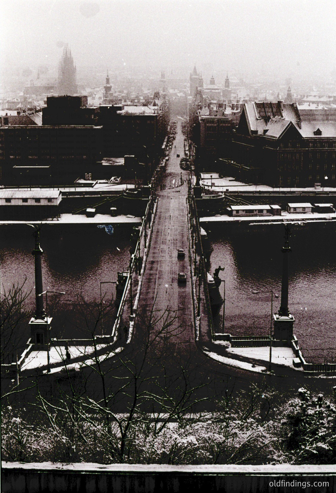 Aerial view of a snow-covered urban canal in winter, likely Budapest. The frozen Danube River splits the cityscape, flanked by historic buildings with gabled roofs. A lone figure stands on a bridge, silhouetted against the misty skyline. Architectural styles suggest late 19th to early 20th century European design.