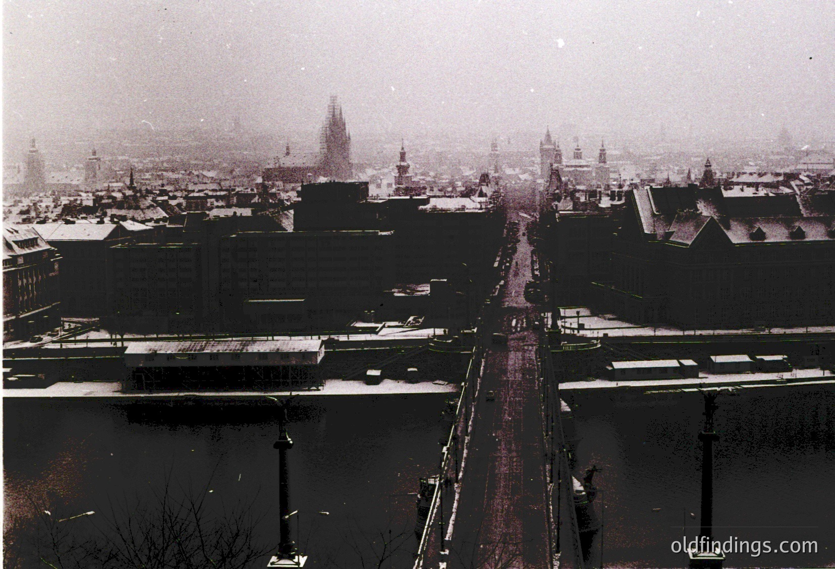 Snow-covered urban street flanked by low-rise buildings, likely European. Prominent church spires and historic architecture in background. Tram tracks and sparse winter traffic visible. Black-and-white sepia tone suggests vintage, possibly 1950s–1970s.