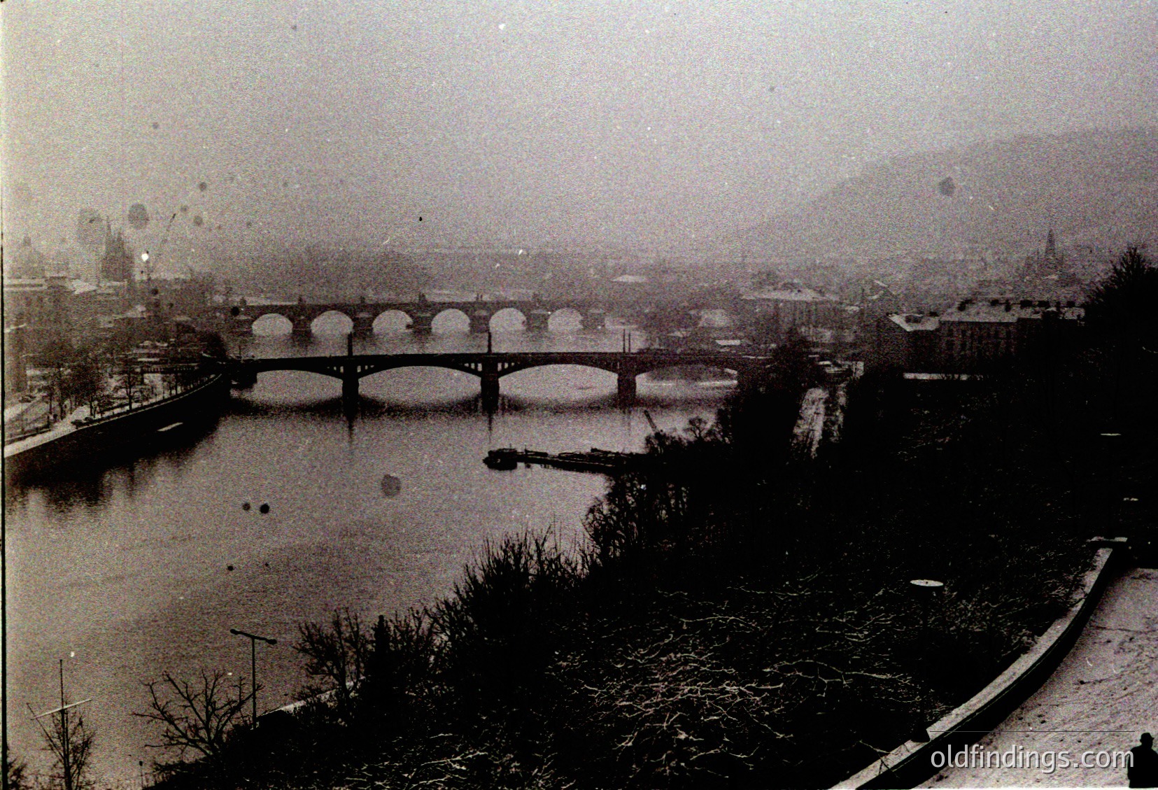 Vintage black-and-white view of a multi-arched stone bridge spanning a river, flanked by sparse winter trees and urban buildings. Fog obscures distant hills, suggesting early 20th-century European cityscape.