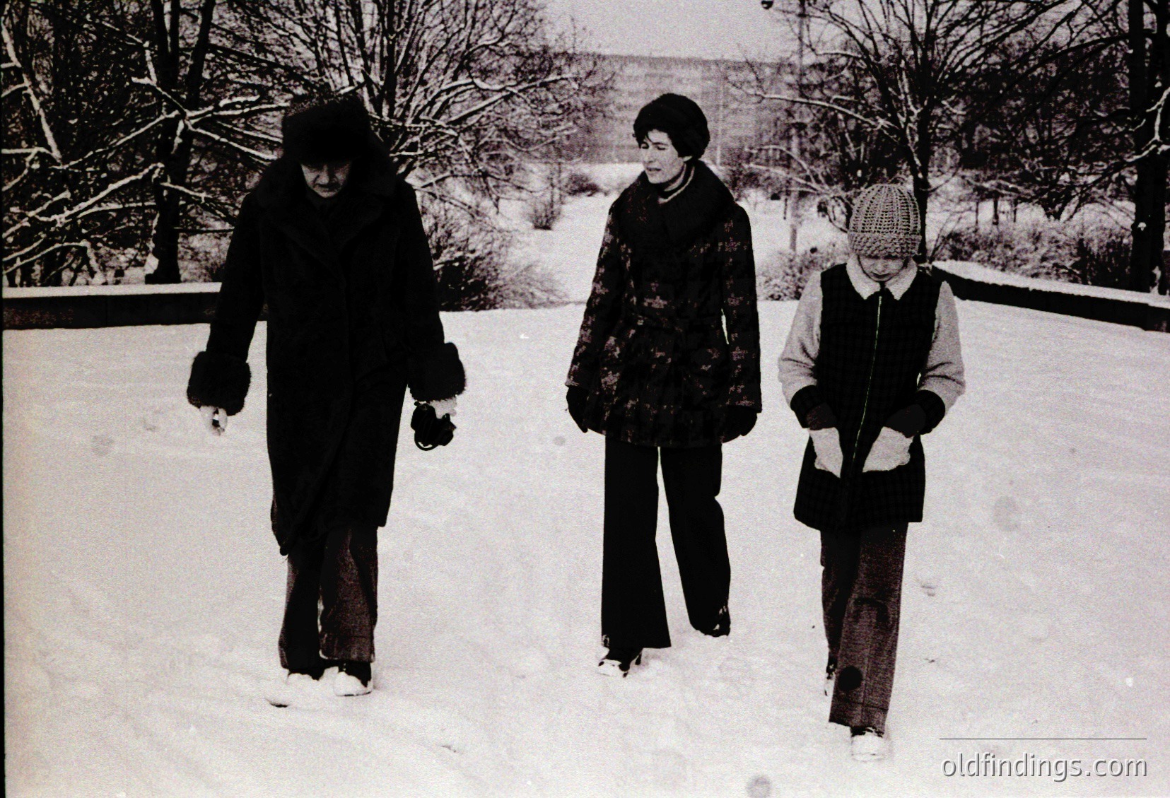 Three individuals in winter attire walk through snow-covered ground, likely a park or residential area. The woman on the left wears a dark coat and hat, the center figure a patterned coat with a scarf, and the child a knitted hat and light jacket. Snow blankets the ground and trees, suggesting a cold season. Style and clothing hint at mid-20th century (1950s–1960s).