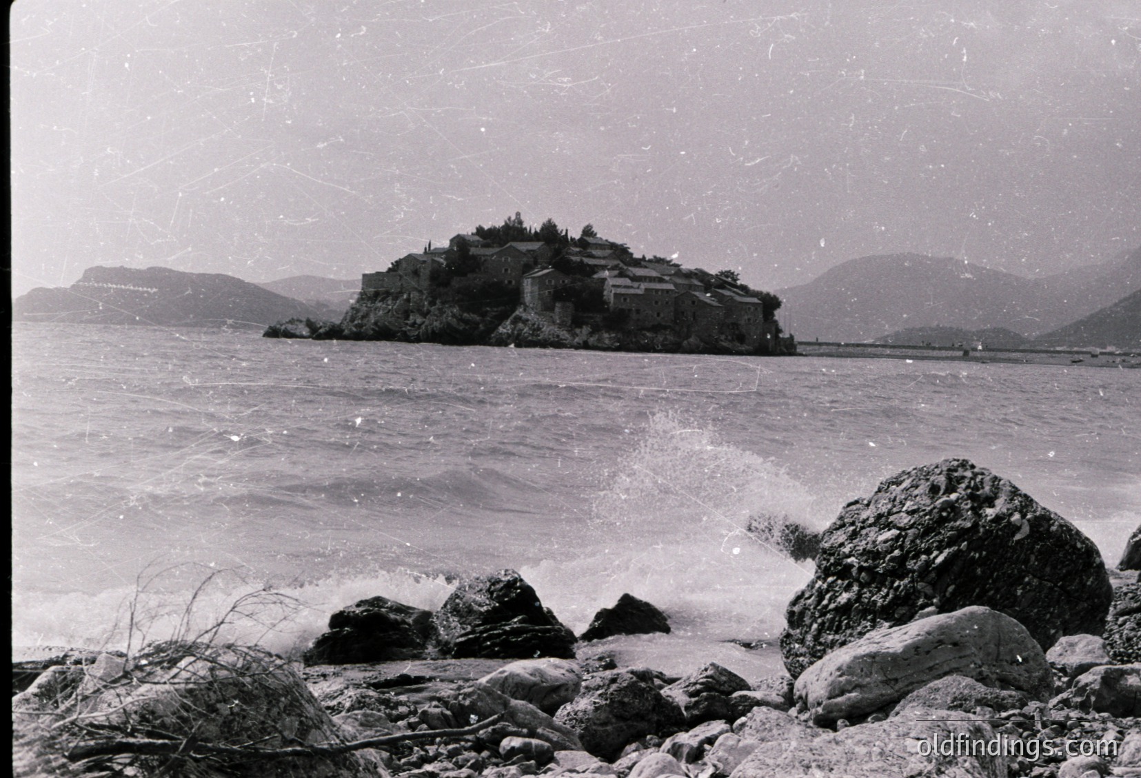Black-and-white coastal scene featuring a fortified island structure atop rugged cliffs, surrounded by choppy waters and rocky shore. Likely a historic military or defensive site, possibly from mid-20th century.