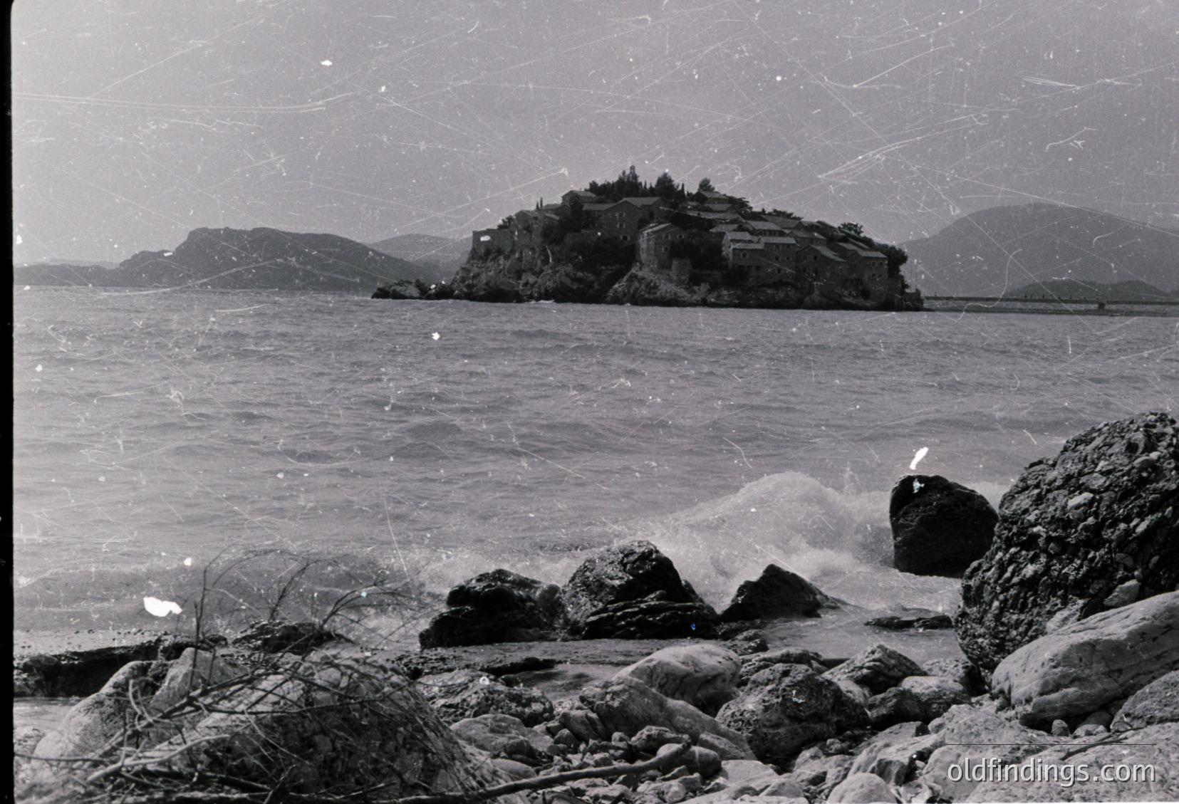 Historic coastal fortress perched on rocky islet, surrounded by turbulent waves. Mid-20th century black-and-white photograph captures rugged Mediterranean architecture and dramatic seascape. Likely or due to similar structures.