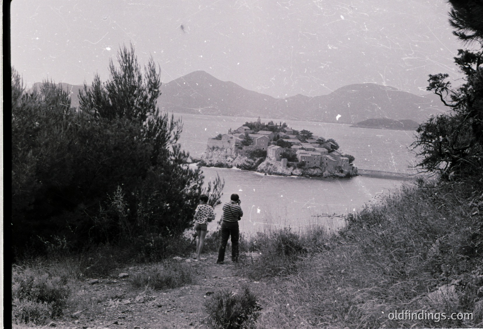 Two figures in winter attire stand on a grassy slope overlooking a rocky island in a frozen lake, framed by snow-laden trees. Mid-20th century monochrome photograph, likely or region. Dramatic winter landscape with rugged terrain and serene water.