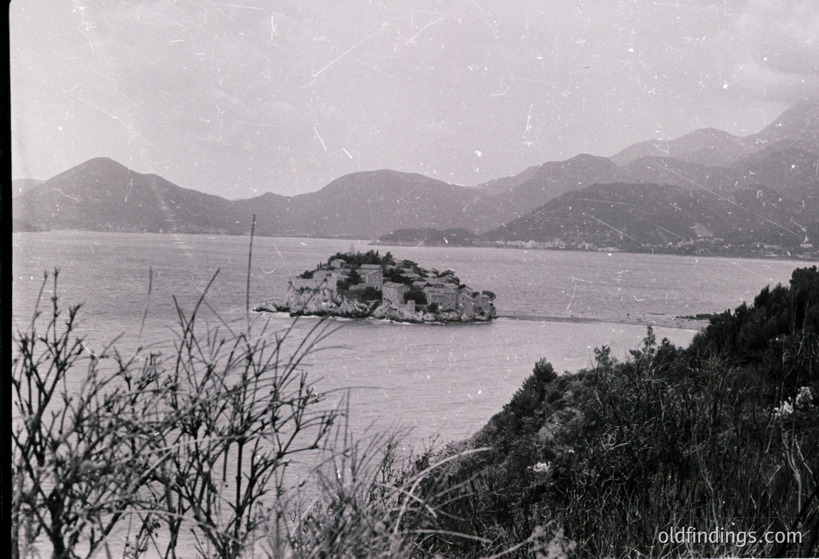 Vintage black-and-white coastal scene featuring a small island fortress surrounded by calm waters, framed by rugged hills. Architectural ruins suggest historical significance, likely from the 19th–early 20th century. Dramatic mountain backdrop enhances the scene’s isolation.