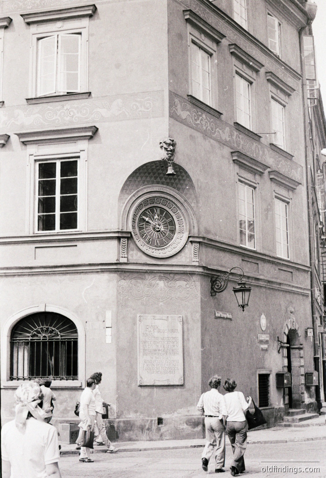 Neoclassical building corner featuring a decorative clock with ornate sunburst design and decorative urn above. Three pedestrians in 1970s-style clothing (bell-bottoms, tunics) walk past. Architectural details include arched windows with iron grilles and wrought-iron street lamp. Likely Eastern European urban setting.