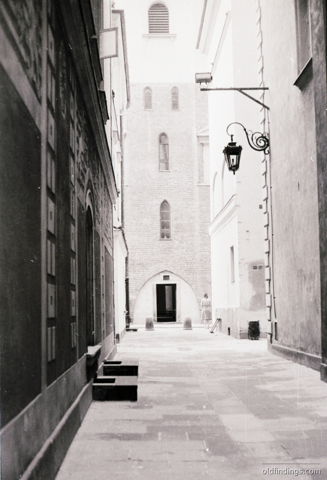 Narrow medieval alleyway with stone-paved path, flanked by tall, weathered buildings. Gothic-style arched doorway at end, flanked by narrow windows with decorative stonework. Classic wrought-iron street lamp affixed to wall. Likely European, 12th–16th century.