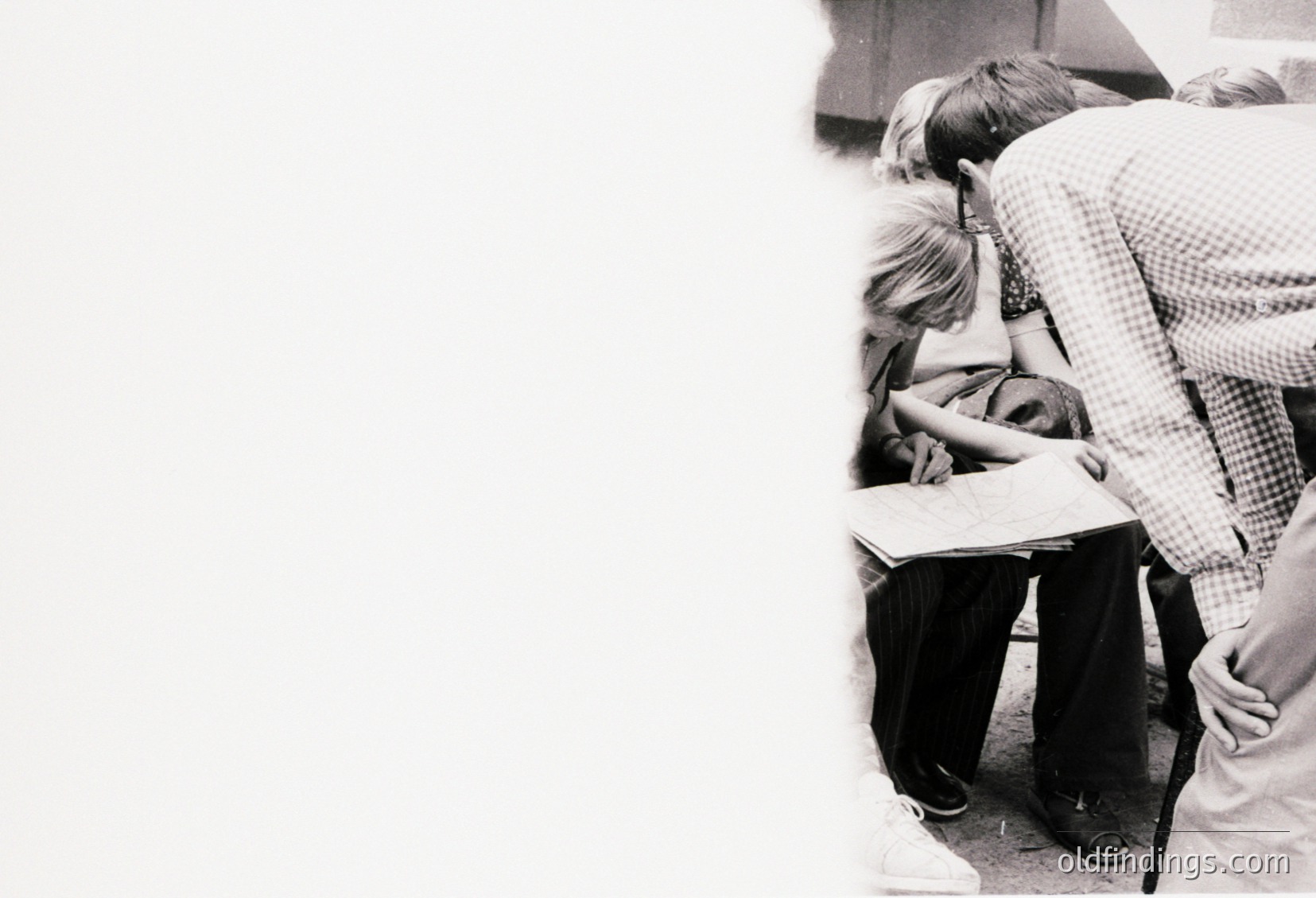 Black-and-white classroom scene showing a teacher (checkered shirt) assisting a seated student at a desk. Another student leans forward, observing. Mid-20th century educational setting, likely 1950s–1960s. Evokes themes of mentorship and learning.