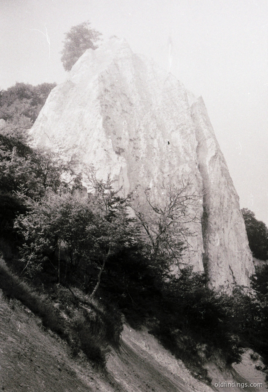 Monolithic rock formation rising sharply from forested terrain, likely limestone. Vegetation includes sparse trees and shrubs on the slope. Dramatic verticality and natural erosion patterns visible.