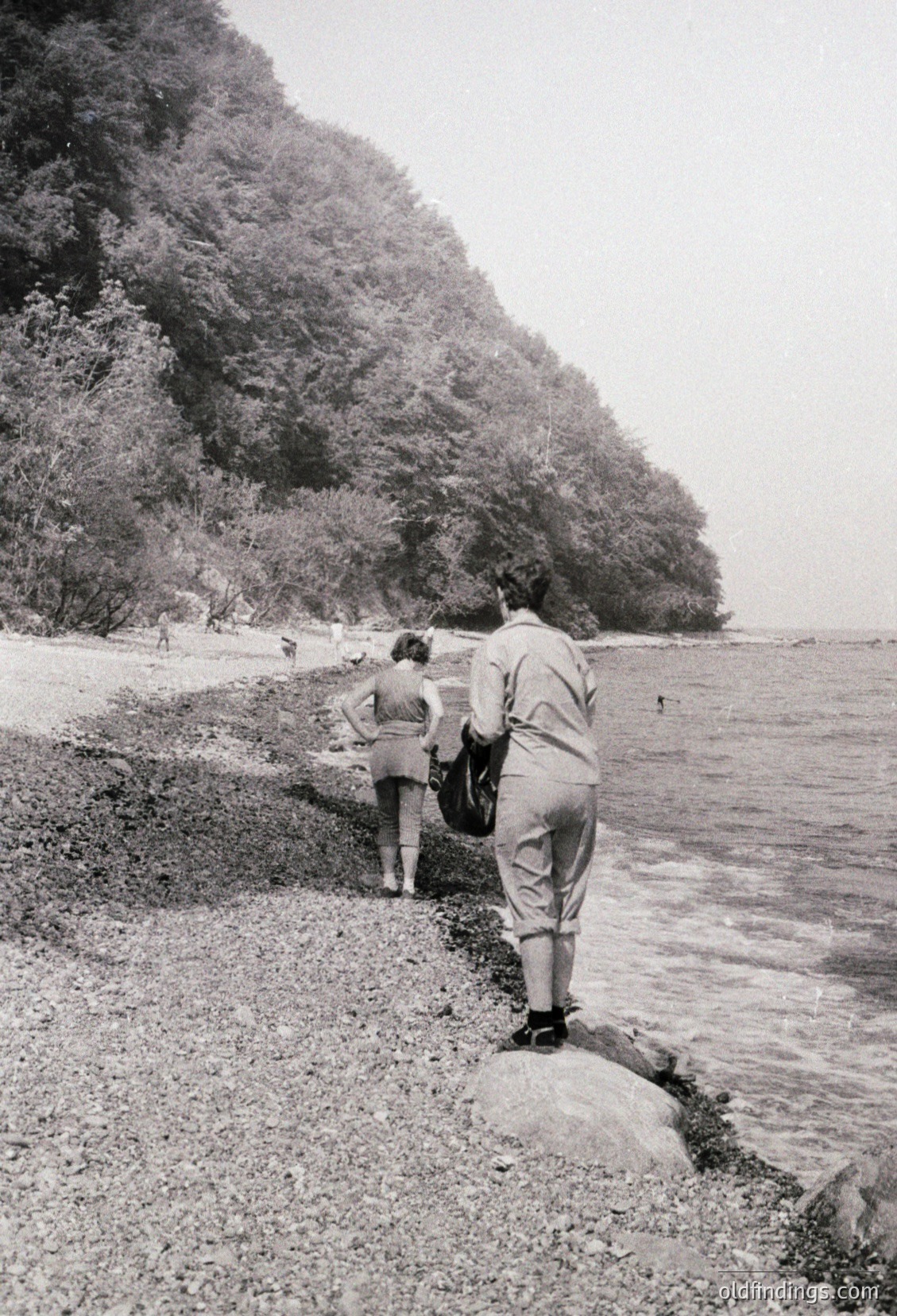 Two figures in mid-20th-century attire (lightweight trousers, short-sleeved shirts) explore a rocky riverside path beside dense foliage. The scene evokes a mid-century outdoor adventure, likely 1950s–1960s.
