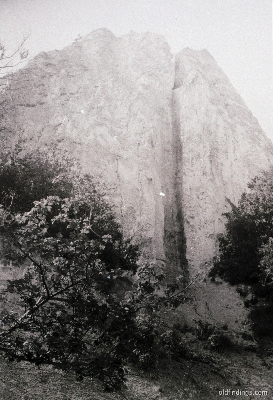 Monolithic limestone cliff with vertical waterfall cascading mid-frame, surrounded by dense foliage. Dramatic natural rock formation likely in a Mediterranean or subtropical region. Black-and-white composition enhances texture and contrast.