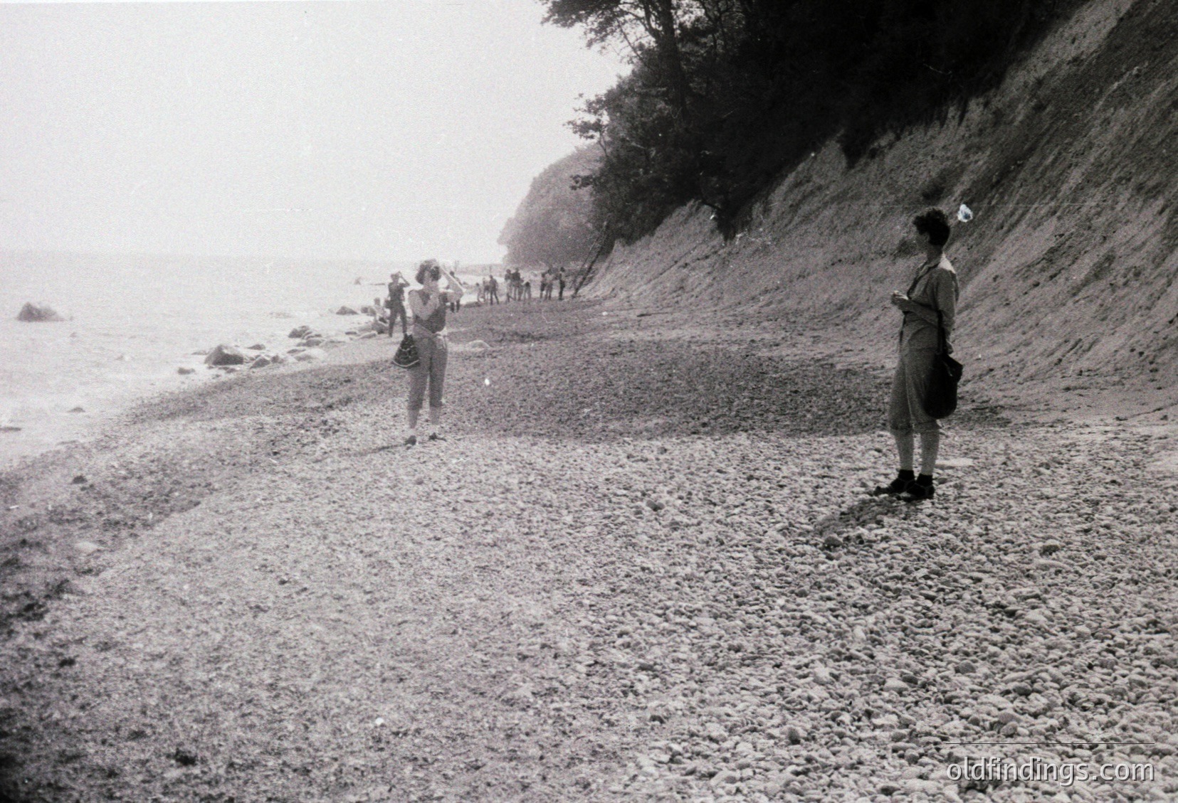 Black-and-white coastal scene showing a gravel path along a rocky cliffside. Two women in mid-20th-century attire (one with a camera, one holding a bag) stand near the path’s edge, overlooking the sea. Crowd of people in the distance follows the path. Mid-century beachside tourism vibe.