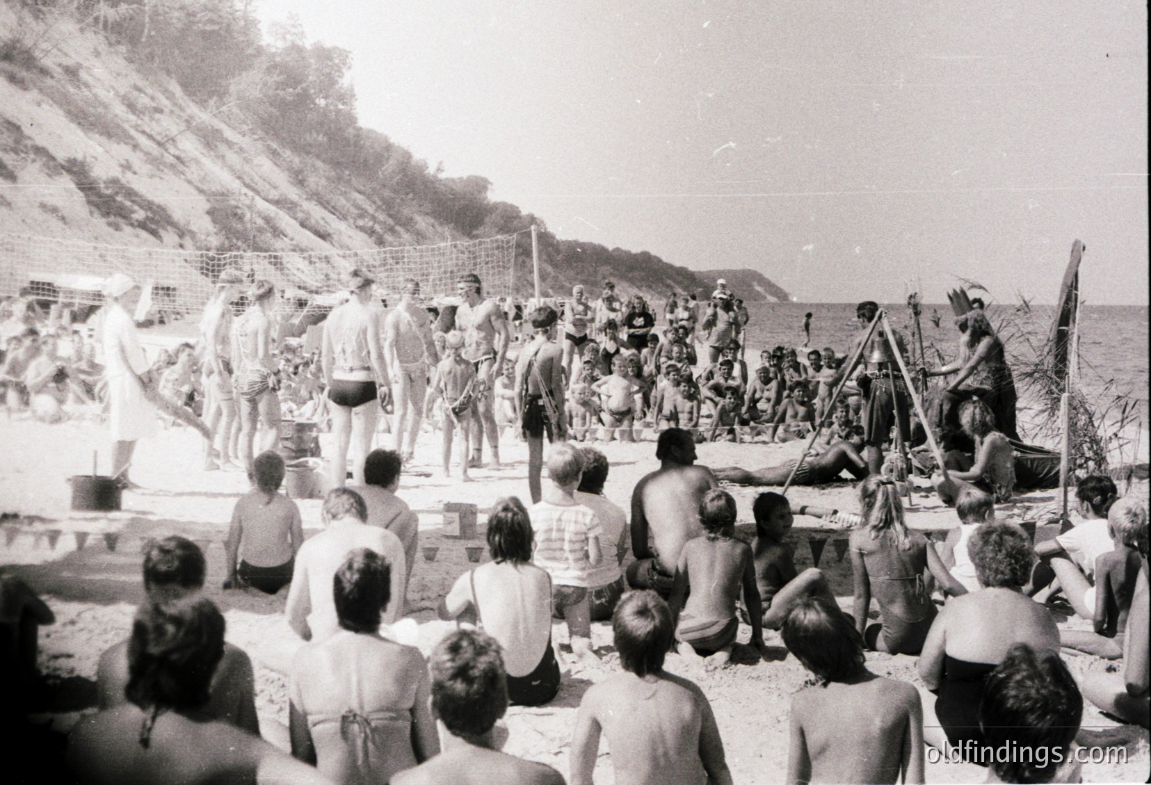 Black-and-white beach scene from mid-20th century, likely 1950s–1960s. Crowded sandy shore with people in swimwear, some seated on blankets, others standing near a makeshift stage. A man in a white shirt and cap directs a group playing a musical instrument. Cliffside and rocky terrain in background.