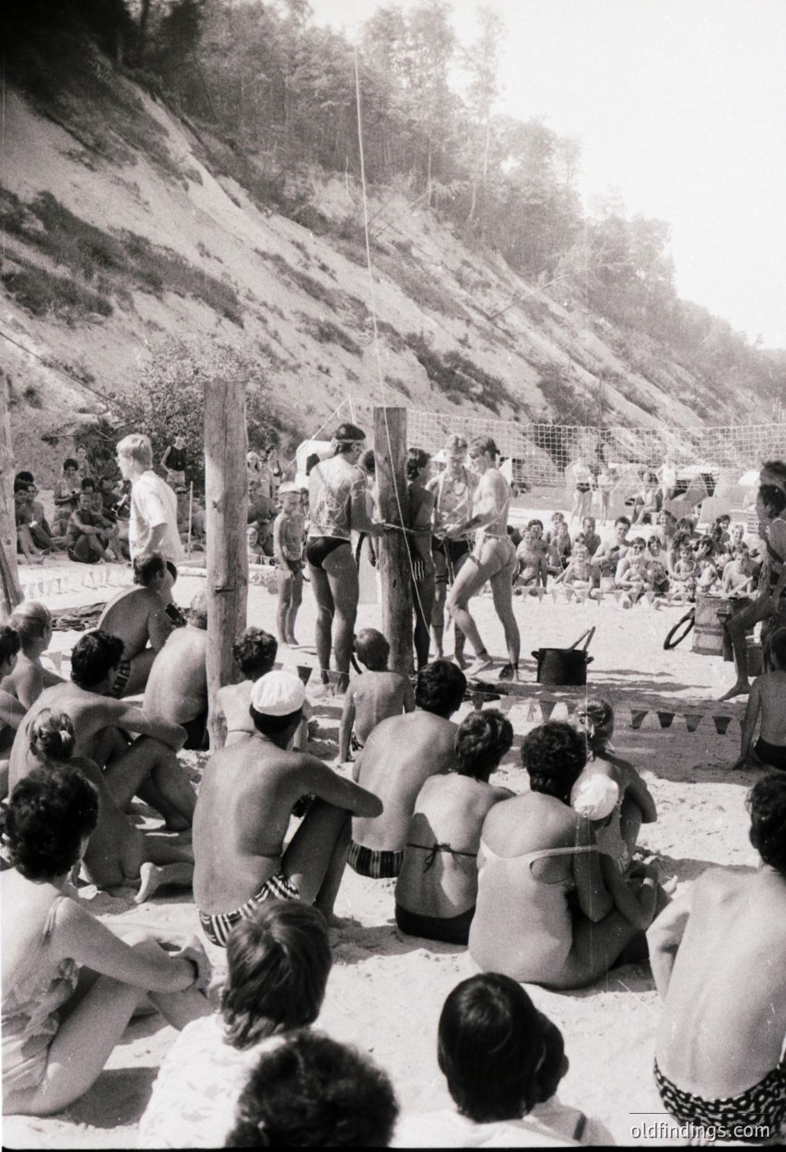 1970s beachside volleyball game with spectators in swimwear, set against rocky cliffs. Crowd seated on sand, players in mid-action. Coastal