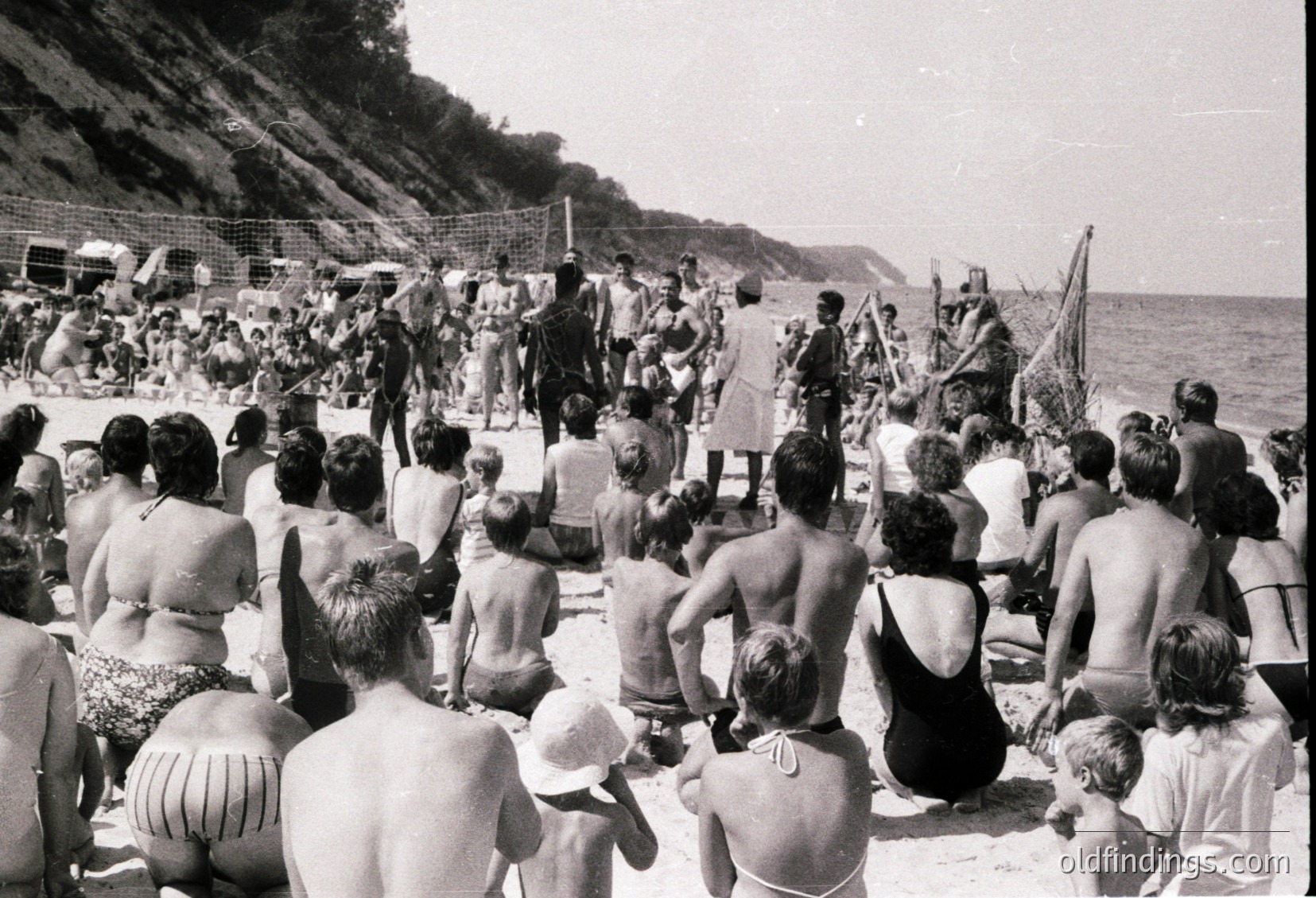 Black-and-white beach scene with a crowd of men, women, and children gathered on sand near a rocky cliffside. Military personnel in uniform stand at attention on a raised platform, likely conducting a ceremony or inspection. Swimmers in swimsuits and hats surround the group, suggesting a mid-20th-century seaside resort atmosphere.
