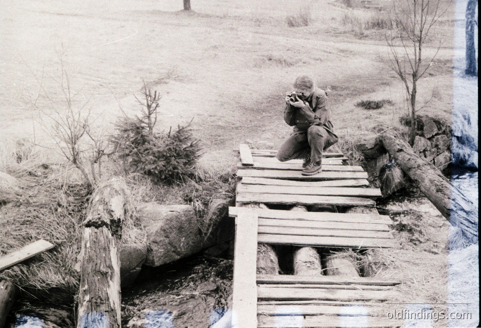 A lone individual sits on a makeshift wooden bridge over a shallow stream in a rural, snow-dusted landscape. The person wears a heavy coat, gloves, and a cap, suggesting cold weather. The bridge is constructed from rough planks and logs, with barrels stacked beneath for support. Snow blankets the ground, trees, and distant fields, indicating a winter setting. The scene evokes a sense of solitude and resilience.