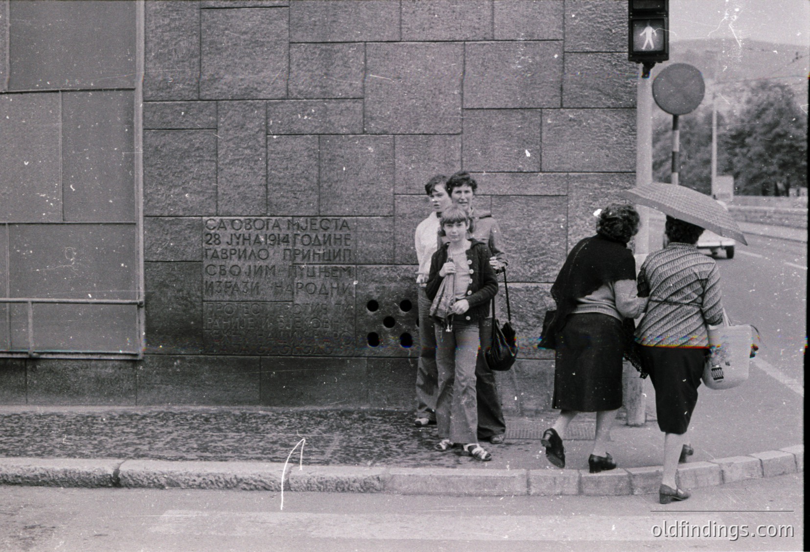 Black-and-white street scene featuring four individuals near a stone monument with Cyrillic text. The monument commemorates a 1941 event ("28 July 1941"). People wear 1970s-era clothing—wide-legged pants, striped shirts, and shoulder bags. One person holds an umbrella, suggesting rain. Urban setting with cobblestone pavement and a pedestrian crossing sign.