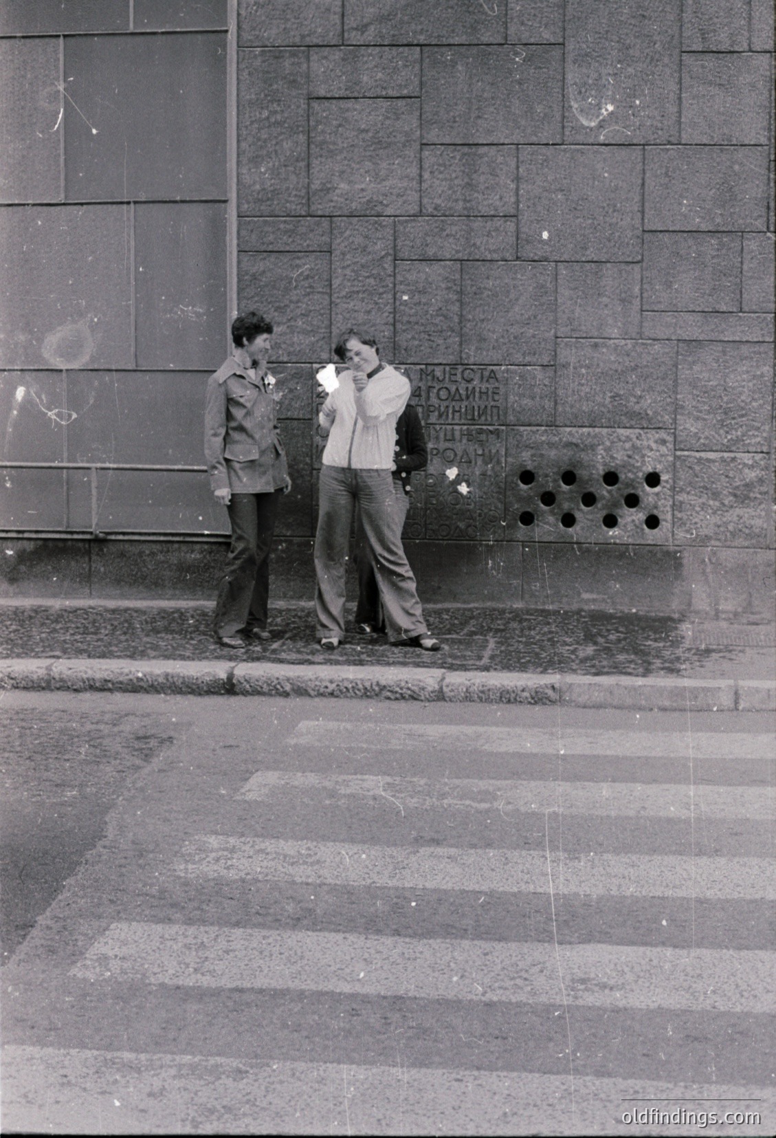 Two young men in 1960s-70s streetwear pose near a Soviet-era concrete wall. One wears a jacket with a pin; Cyrillic text ("КУЛТУРНЫЙ ОТДЫХ") suggests Eastern Bloc location. Urban street scene with patterned pavement.