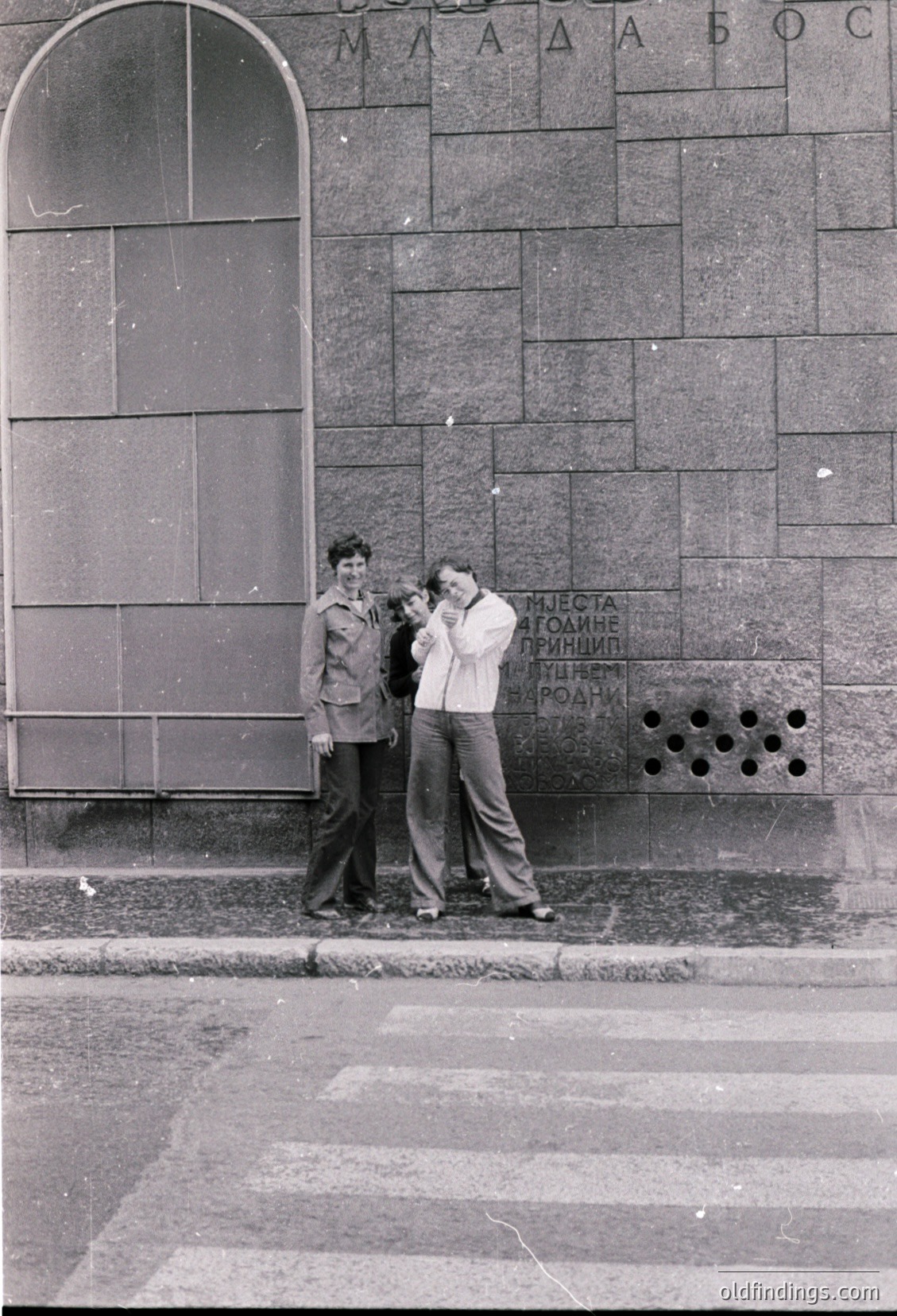 Two men pose against a stone wall with Cyrillic text in 1960s-70s Sofia, Bulgaria. The wall bears Soviet-era inscriptions. One man wears a jacket with shoulder epaulettes; the other sports a white shirt and jeans. Urban street scene with patterned pavement.