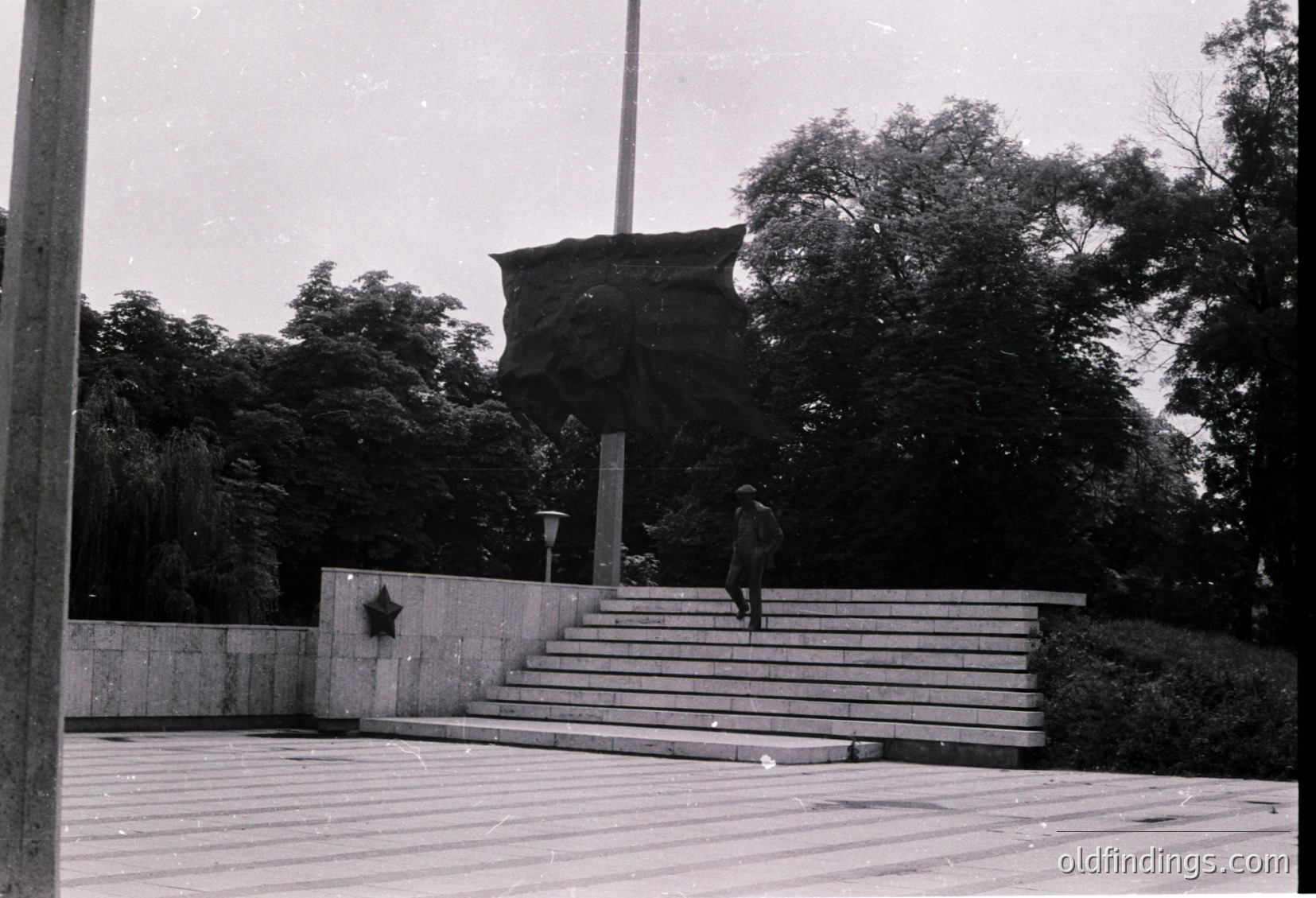 Monumental Soviet-era memorial with a hammer-and-sickle emblem on a concrete base, flanked by a large abstract stone relief. A lone figure in a hat descends the wide concrete steps. Mid-20th century urban park setting, likely Eastern Bloc.