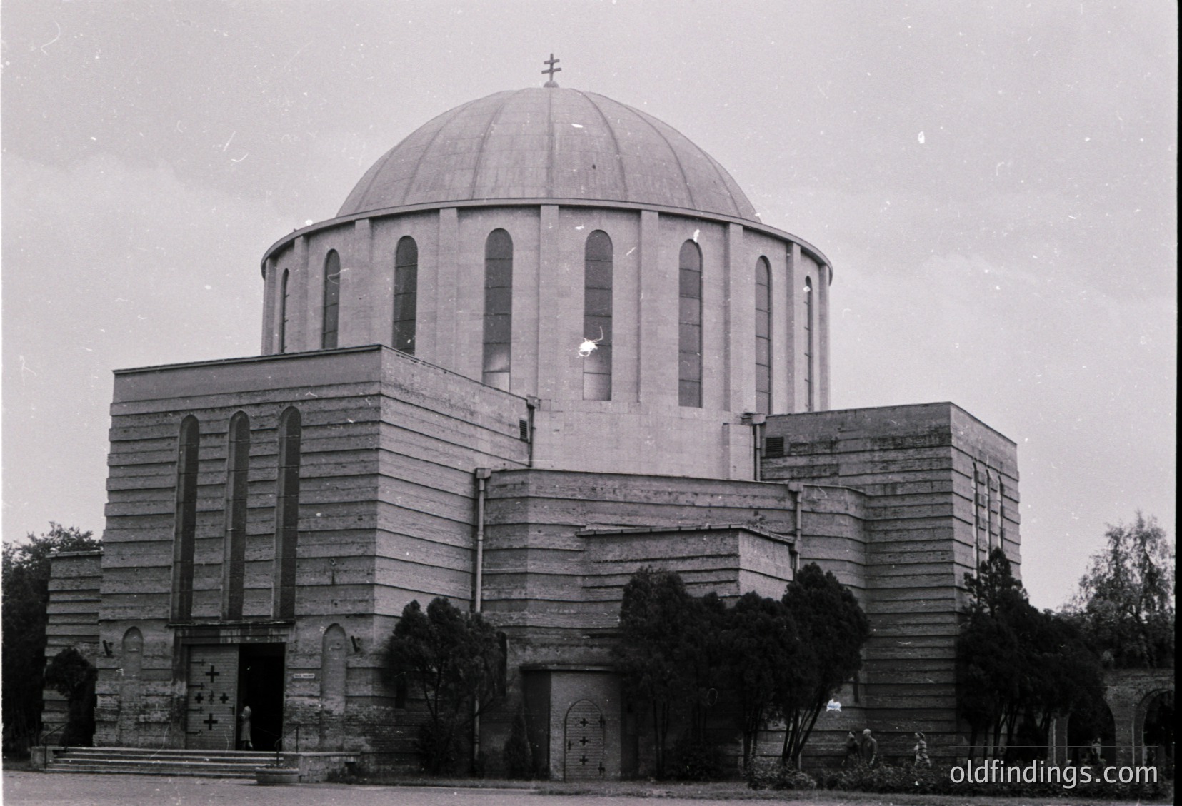 Mid-century modernist church with cylindrical drum dome and brutalist concrete façade. Cross-topped central dome flanked by rectangular extensions. Architectural details include arched windows and a prominent entrance with cross symbol. Likely Eastern European, 1960s-1970s.