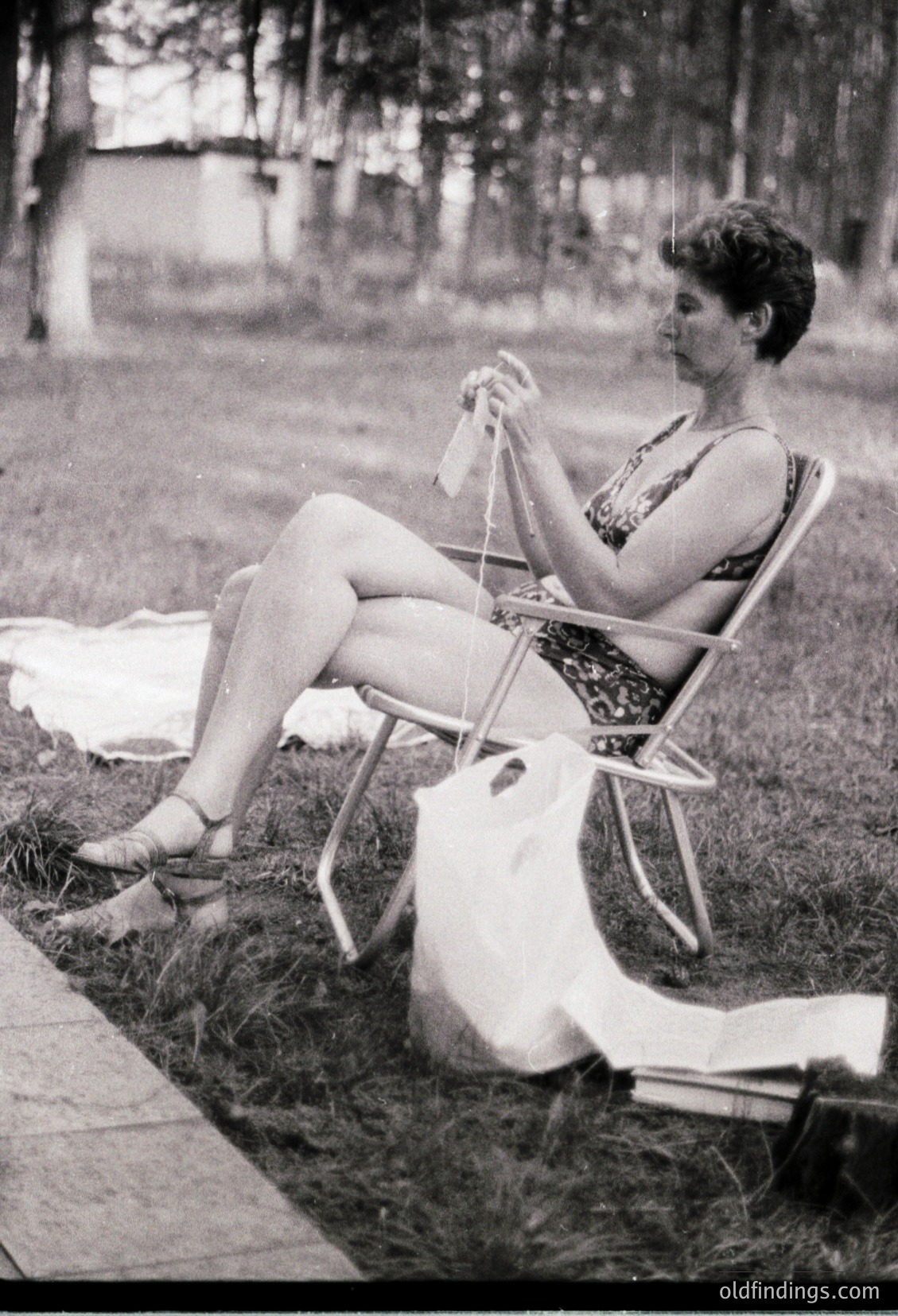 Mid-century outdoor scene: Woman in sleeveless dress and sandals knits beside a picnic blanket on grass, seated on a metal chair. Wooded park background suggests 1950s–1960s leisure culture.