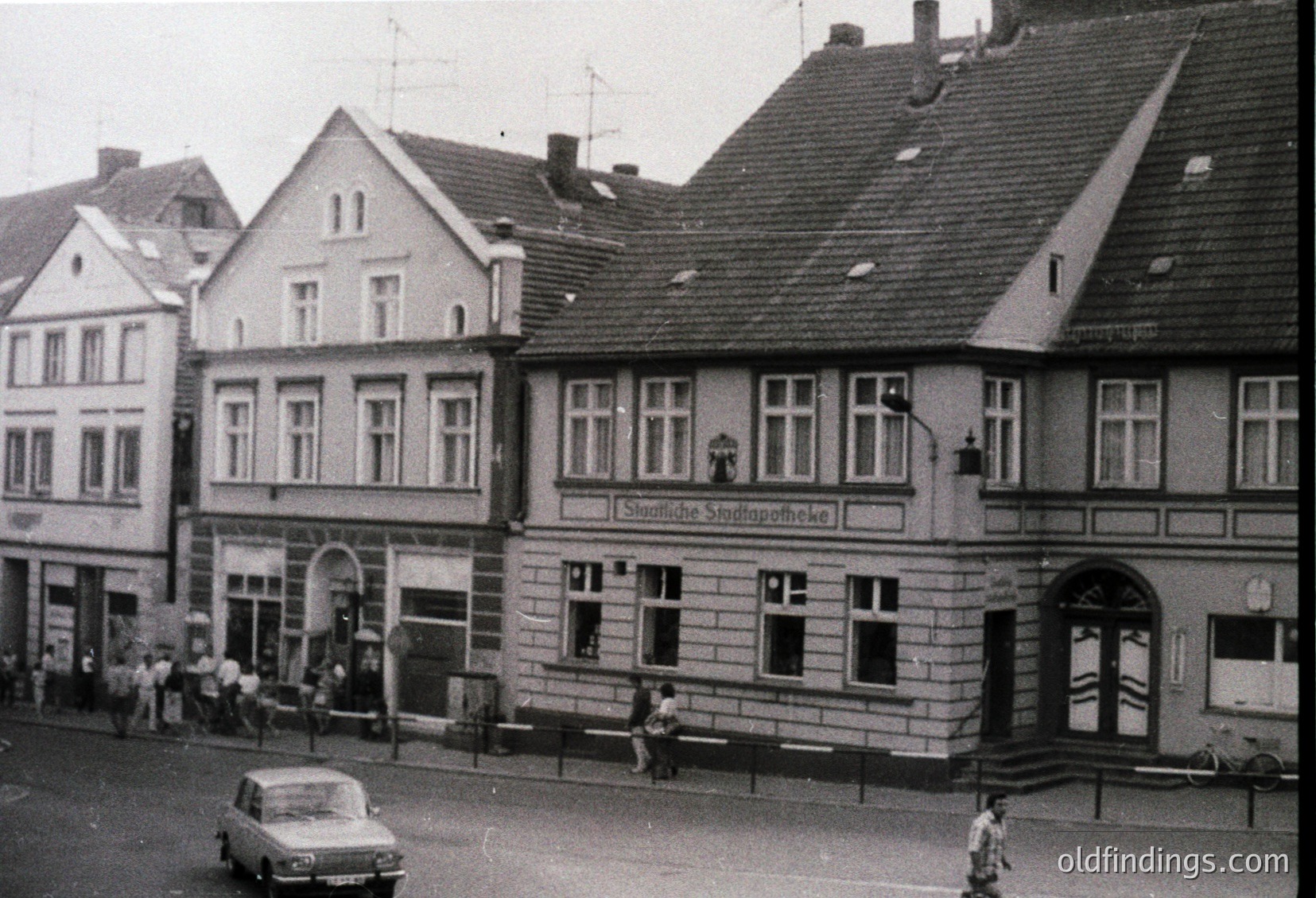 Mid-century European street corner featuring three-story brick buildings with gabled roofs and symmetrical windows. The central building displays a sign reading *"Städtische Sparkasse"* (municipal savings bank) in Gothic script, indicating a 1950s–1970s German-speaking locale. A vintage car and pedestrians suggest a quiet urban scene.