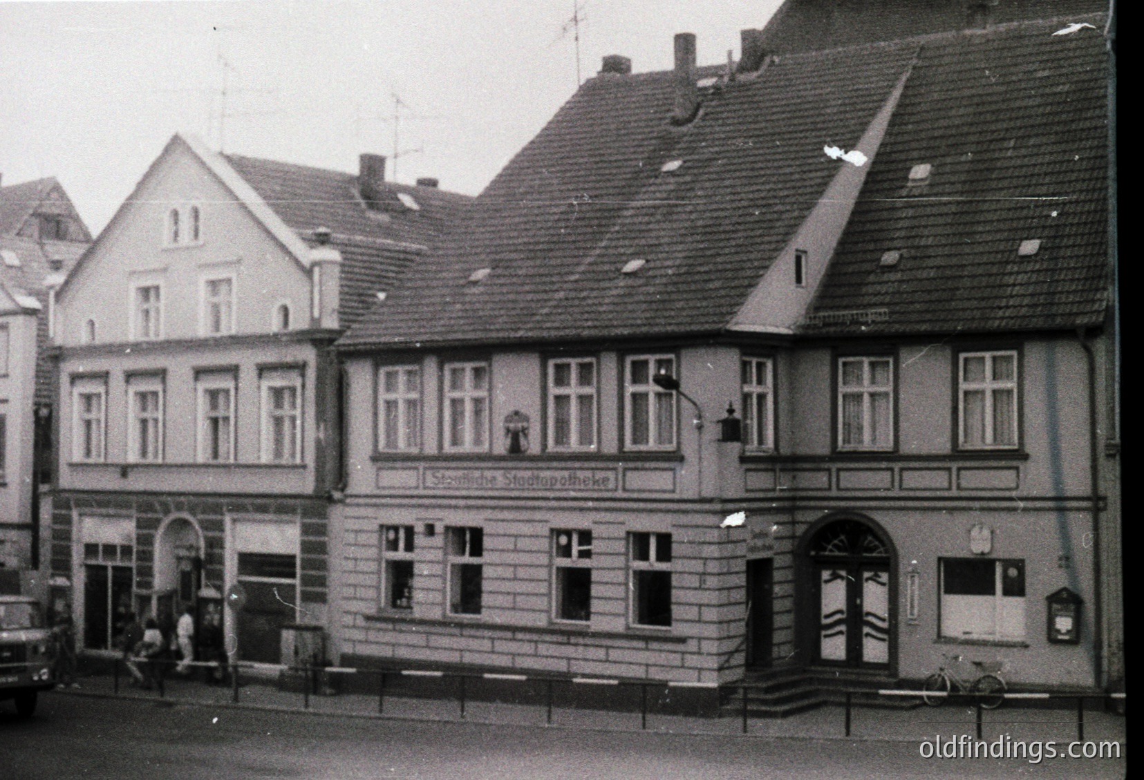 Three-story corner building in classic German urban architecture, featuring gabled roofs, symmetrical windows, and a sign reading *"Städtische Stadtspotheke"* (municipal pharmacy). Black-and-white, likely 1950s–1960s. Street-level storefronts with arched entrances and wrought-iron railings.