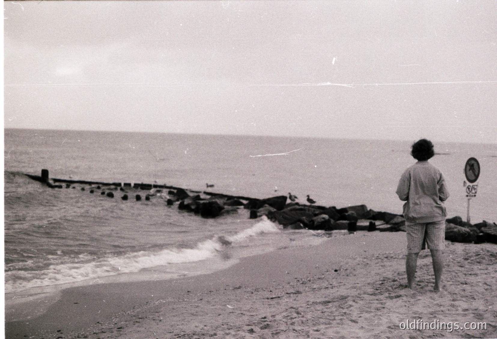 Child stands on a seaside promenade with concrete pier, waves crashing against stone breakwater. Overcast sky suggests mid-20th century coastal scene, likely Eastern Bloc era.