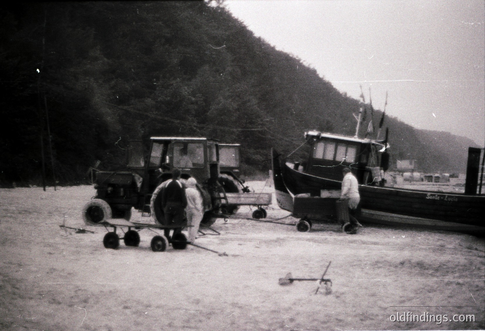 Mid-20th century coastal scene: two men inspecting a vintage tractor and a wooden fishing boat on sandy shore. Hilly, forested background suggests a sheltered harbor.