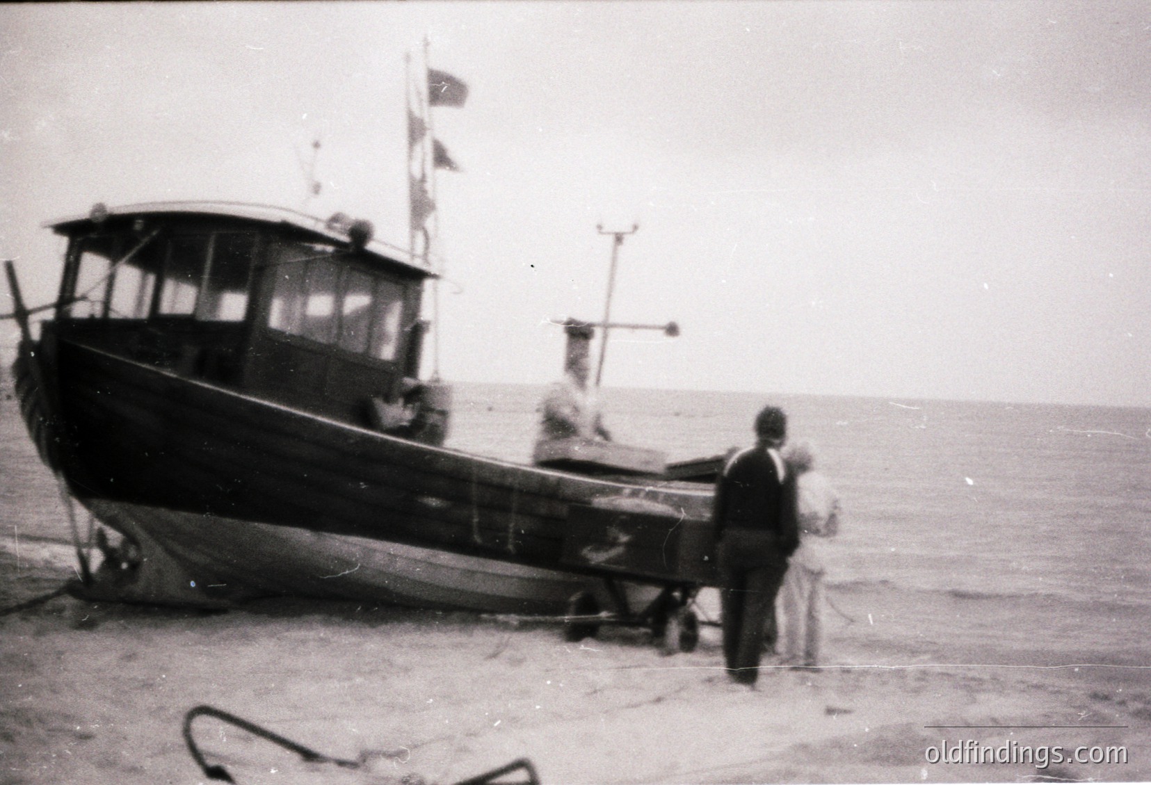 Black-and-white mid-20th century fishing boat on a sandy shore, likely Eastern European coastal region. Two men in dark suits and a woman in a light dress stand near the vessel’s bow, which features a small cabin and flagpole. Boat’s hull shows signs of wear, suggesting active use.
