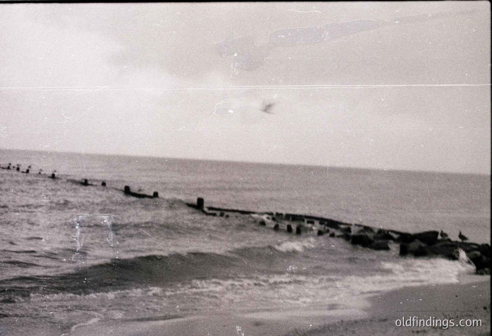 Black-and-white coastal scene featuring a weathered wooden pier extending into choppy waters. The structure appears aged, with visible gaps and erosion. Seagulls perch along the pier’s edge, while waves crash against the shore. Likely mid-20th century, possibly , due to photographic style and pier construction.