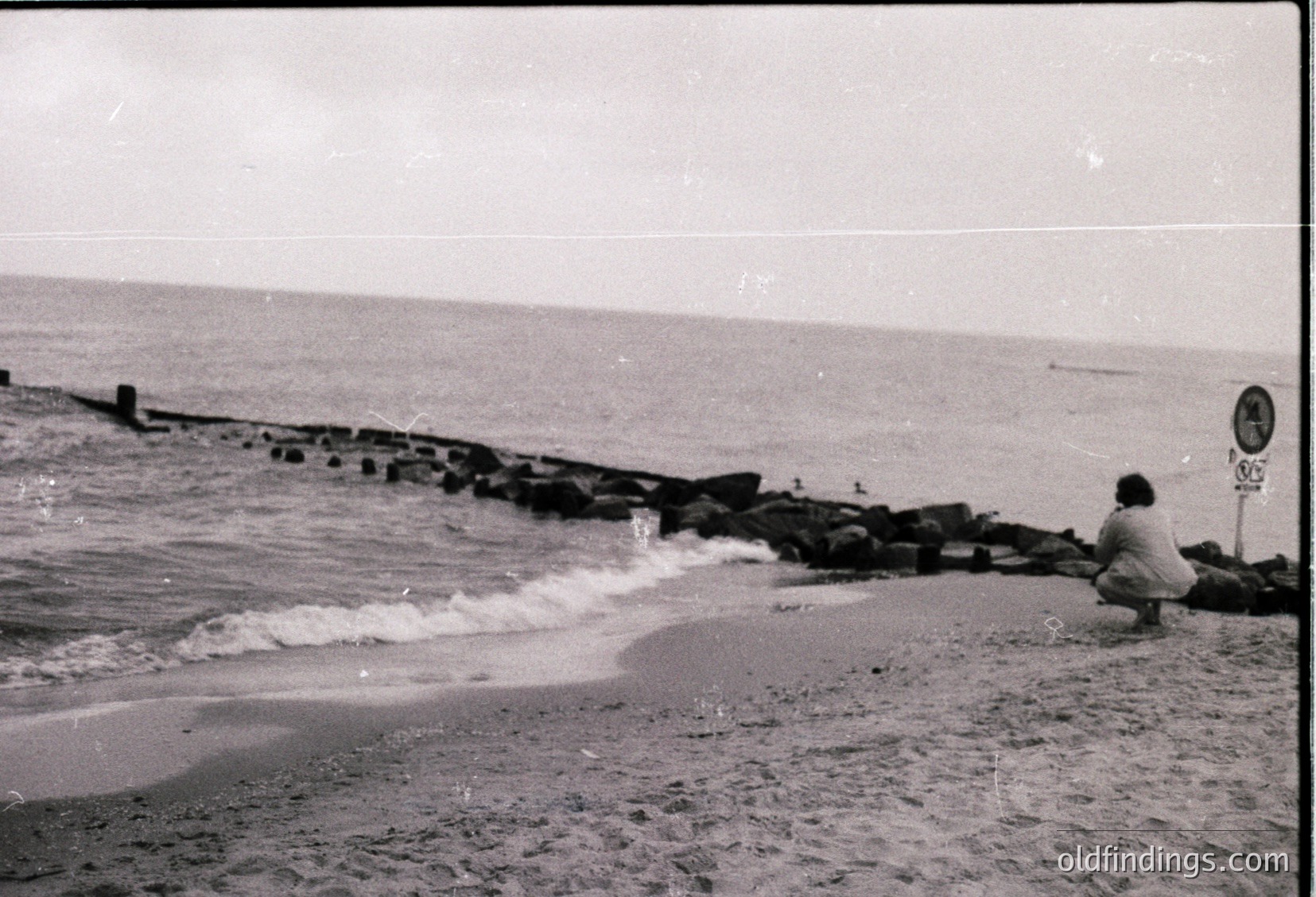 Black-and-white seaside scene featuring a lone figure sitting on a concrete pier by crashing waves, likely mid-20th century. Concrete breakwater and wooden posts line the shore, with a signpost in the background. Evokes midcentury coastal architecture and solitude.
