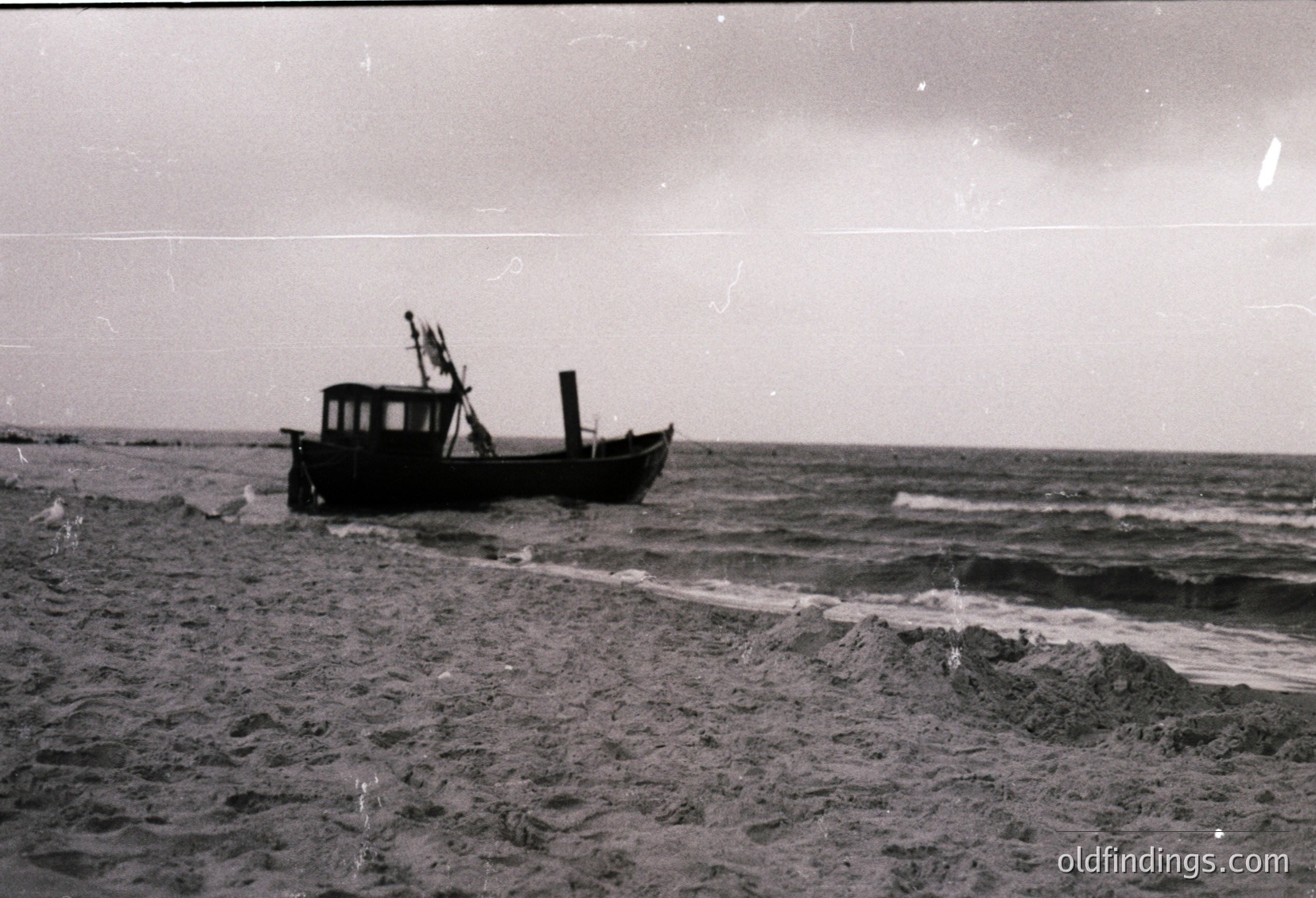 Vintage black-and-white image of a small wooden fishing boat grounded on a sandy shore, waves crashing nearby. Open sea and cloudy sky in background. Likely mid-20th century coastal scene.