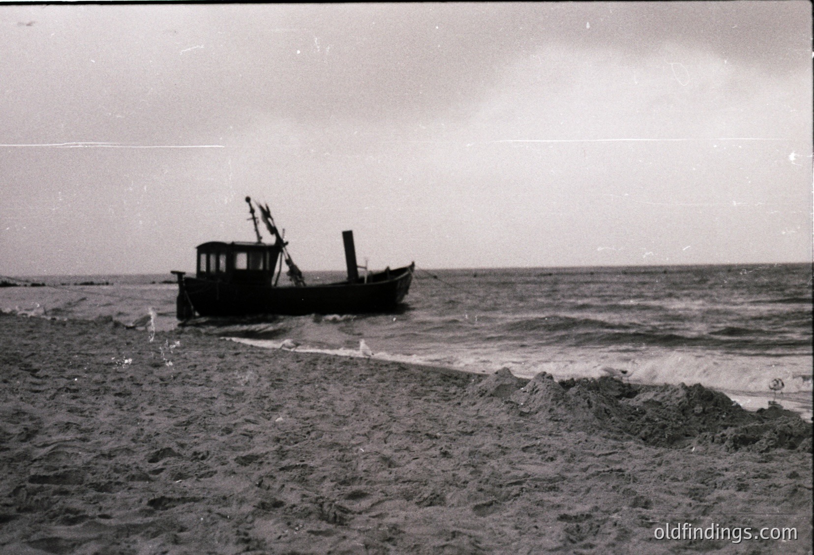 Vintage black-and-white photo of a small wooden fishing vessel grounded on a sandy shore, waves crashing nearby. Mid-20th century coastal scene, likely Eastern European.